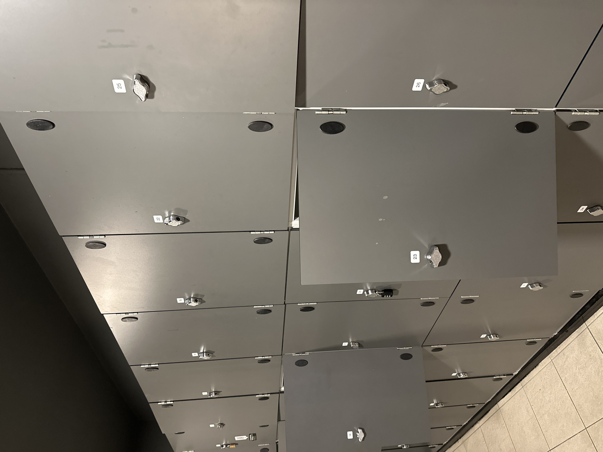 The image displays a section of grey metal lockers arranged in a grid. The lockers have round, black knobs or push-button locks. Some locker doors are ajar, revealing the interior. Several lockers are labeled with white rectangular stickers displaying numbers, such as 14, 22, 23, 25, and 26. The lockers are positioned in a room with a dark ceiling and tiled flooring. The perspective suggests an indoor environment, likely a public facility such as a gym or changing room. There are no people visible in the image. The overall scene is orderly and functional. This image does not provide any information about its location in Carlow, Ireland.