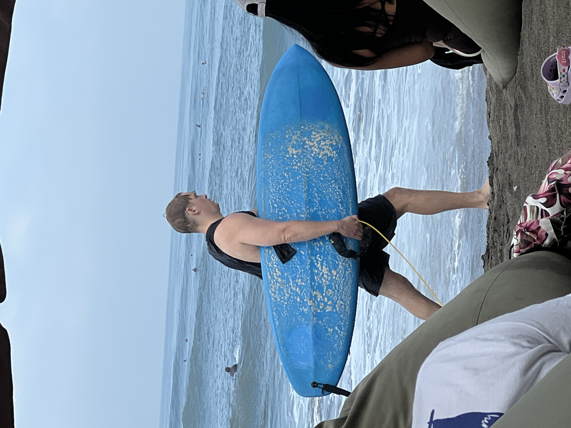 In North Kuta, Indonesia, a light-skinned man with short blonde hair, wearing a black tank top and black shorts, carries a bright blue surfboard with white speckles. He is walking from left to right across the frame, with the surfboard held horizontally and in front of him. He has a yellow leash attached to the surfboard, which trails behind him. He has a backpack on his shoulders. The man is walking on a dark sand beach, with waves of the ocean reaching the shore. In the background, several people are visible in the water, some of whom appear to be surfing. In the foreground, to the left and right, parts of beanbag chairs are visible, suggesting a seating area on the beach. One of the beanbag chairs on the right has a portion of a person with dark hair sitting on it. A child's pink Croc shoe is on the sand in the lower right corner. The sky is overcast, and the ocean appears calm with gentle waves.