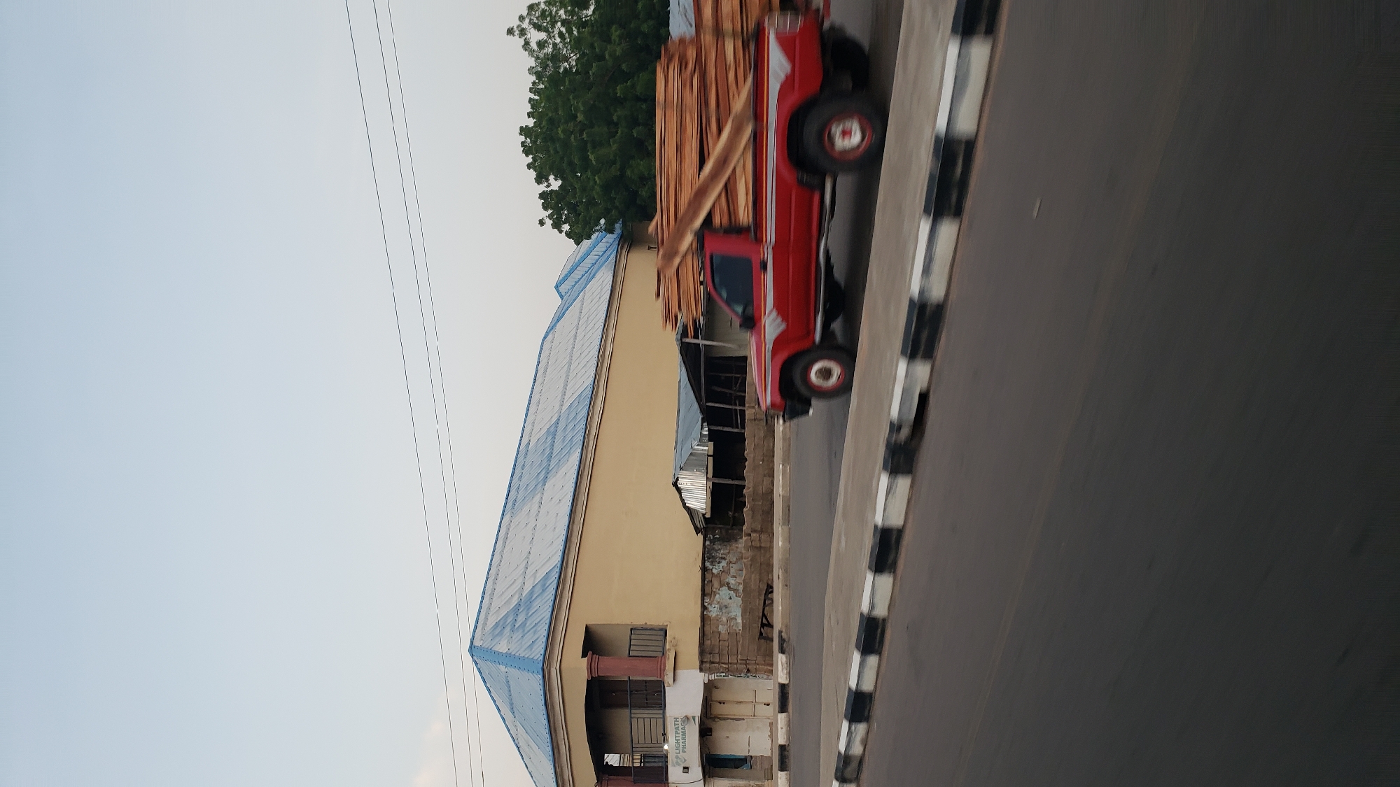 A red pickup truck laden with stacks of lumber is driving on a road in Jalingo, Nigeria. The truck is moving from left to right across the frame. The lumber is piled high in the truck bed and extends out over the cab. The truck has silver accents on its sides. In the background, a two-story building with a blue corrugated metal roof is visible. To the left of the building is a sign that reads "LIGHTPATH PHARMACY". Adjacent to the building are some open-air structures with corrugated metal roofs. A line of trees is visible behind these structures. The road has a striped curb with alternating black and white sections. The sky is overcast.