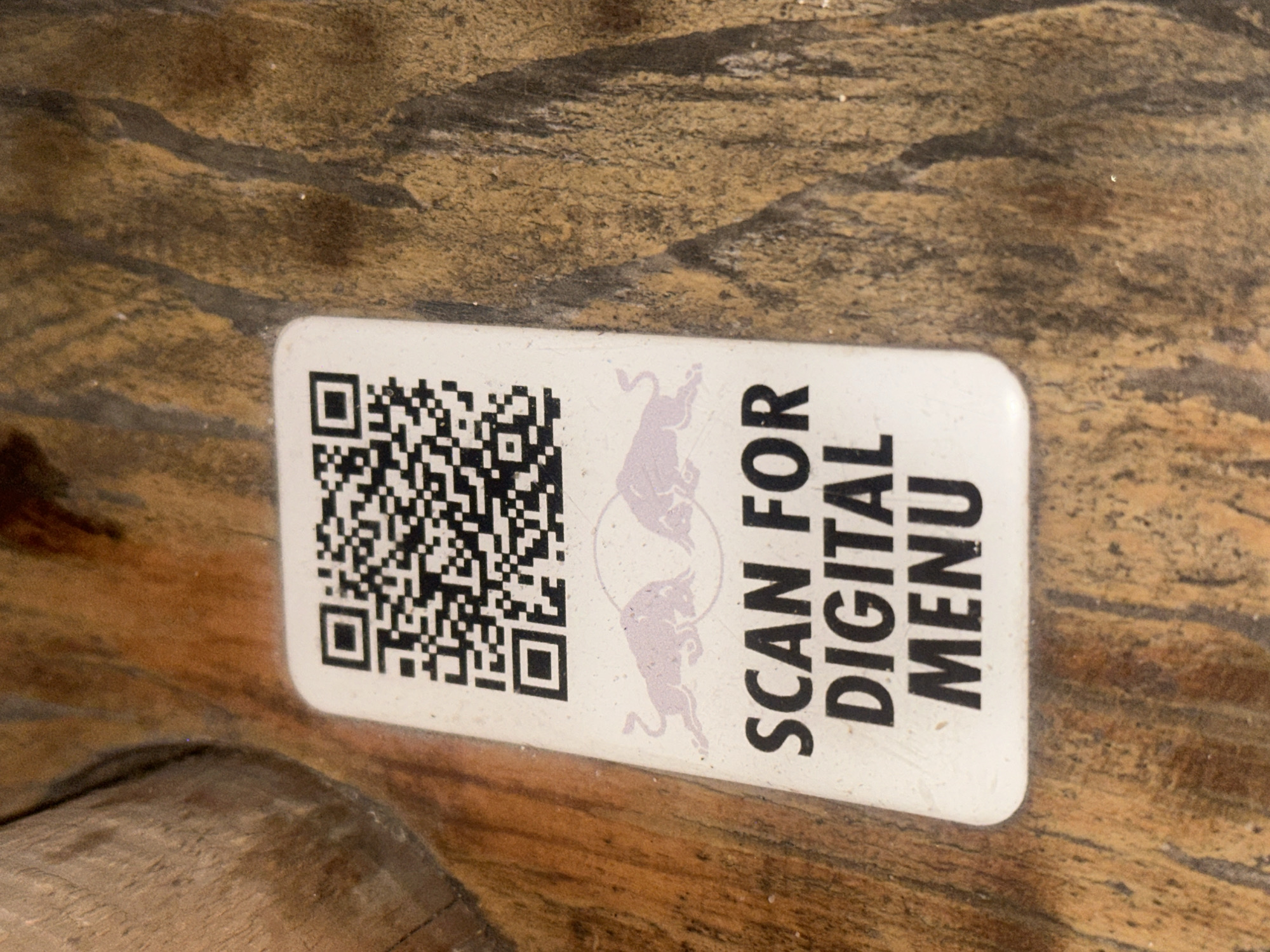 A white, rectangular sticker with rounded corners is affixed to a rustic, reddish-brown wooden surface, likely a tabletop or bar, within an establishment in the city of Unknown, Unknown. The sticker features a black and white QR code at its upper portion. Below the QR code is a stylized graphic of two light purple bulls facing each other, characteristic of the Red Bull brand. The bottom section of the sticker displays the black, bold, sans-serif text 'SCAN FOR DIGITAL MENU' across three lines. The wooden surface exhibits prominent grain patterns, darker streaks, and a somewhat worn or uneven texture, with specks of dust visible. To the far left, a smoother, lighter brown wooden element is partially visible, contrasting with the main surface.