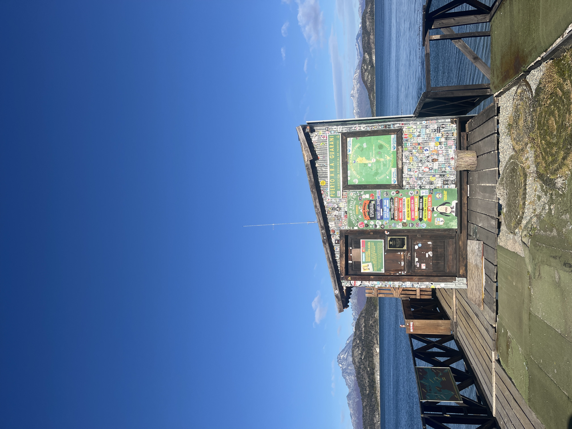 The image shows a small, rustic building on a wooden platform extending over a body of water, likely a lake or ocean. The building is covered in numerous stickers and signs, including a large green sign with a map and text that reads "PUERTO A.R.A. GUARANI," "CORREO UNIDAD POSTAL," and "FIN DEL MUNDO".
Other signs indicate distances to various cities like Ushuaia, Buenos Aires, and places like the South Pole and Antarctica. The building appears to be a post office or a similar station, given the mentions of "CORREO" (post office) and "ESTAFETERO" (postman).
The surrounding environment features mountains in the background and a clear blue sky with some clouds. The water is a dark blue color. The scene evokes the feeling of a remote, possibly tourist-visited location. The image composition is vertical, with a large portion of the sky dominating the left side of the frame.