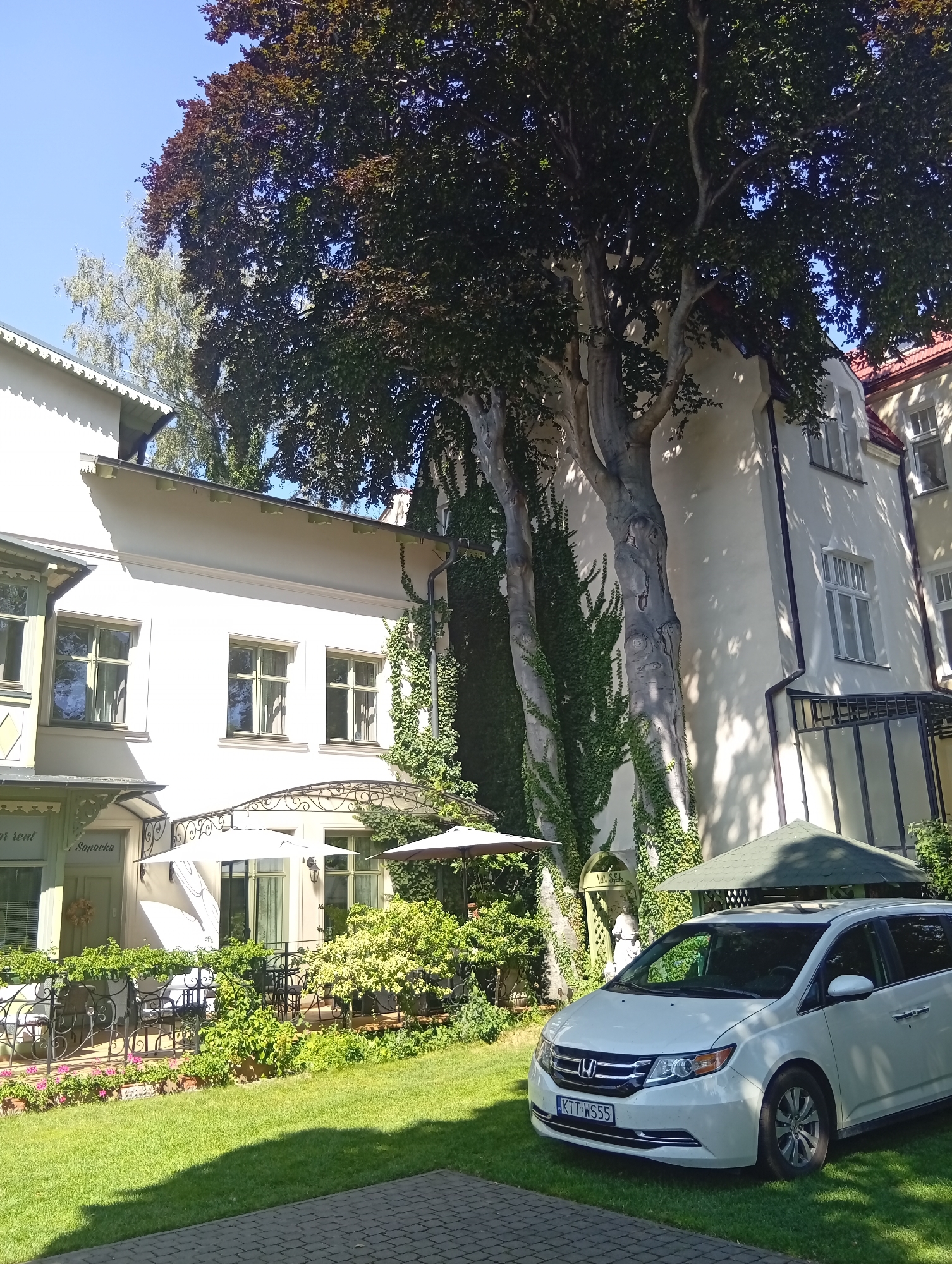 The image shows a white minivan parked on a green lawn in front of a building. The building appears to be a house with a white facade and red-tiled roofs. There are trees with dark leaves and ivy growing on the side of the building. In the foreground, there is a paved walkway. The license plate of the minivan is visible and reads "KTT WS55". Based on the context of the building's architectural style and the presence of Polish license plates, the location is likely Sopot, Poland. The scene depicts a residential or commercial property on a sunny day, with no people visible.