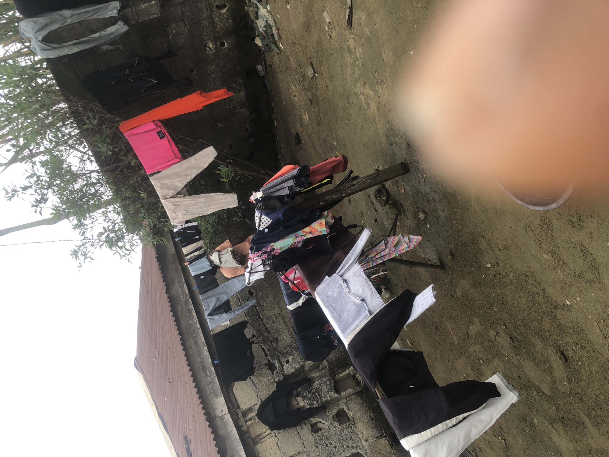 The image depicts an outdoor scene in Ado, Nigeria, featuring a clothesline strung with various items of laundry. The clothesline is suspended between a weathered brick wall and a makeshift wooden support. The laundry includes a mix of garments such as trousers, shirts, and other clothing items, some of which are brightly colored. Behind the clothesline, a corrugated iron roof of a building is visible, and lush green foliage can be seen growing in the background. The ground appears to be dirt, with some scattered debris, suggesting a somewhat unkempt environment. In the foreground, a blurred portion of what appears to be a person's hand or finger is obscuring part of the scene, suggesting the photograph was taken by someone present.