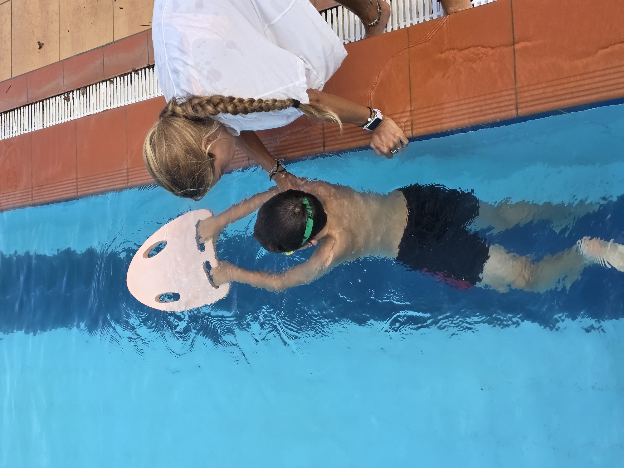 A woman in a white dress and a blonde braid is at the edge of a swimming pool, helping a child swim. The child is holding a pink kickboard and wearing dark swim trunks and green goggles. The pool has orange tile trim. The location is likely Phu Thuy Ward, Vietnam.