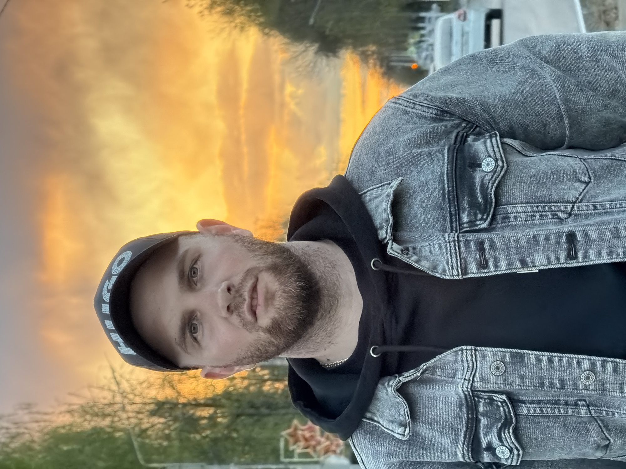 A medium shot captures a man outdoors at sunset. He wears a black hoodie under a gray denim jacket and a black baseball cap. The background shows trees and buildings. The location is potentially the city of Chaikovsky, Russia.