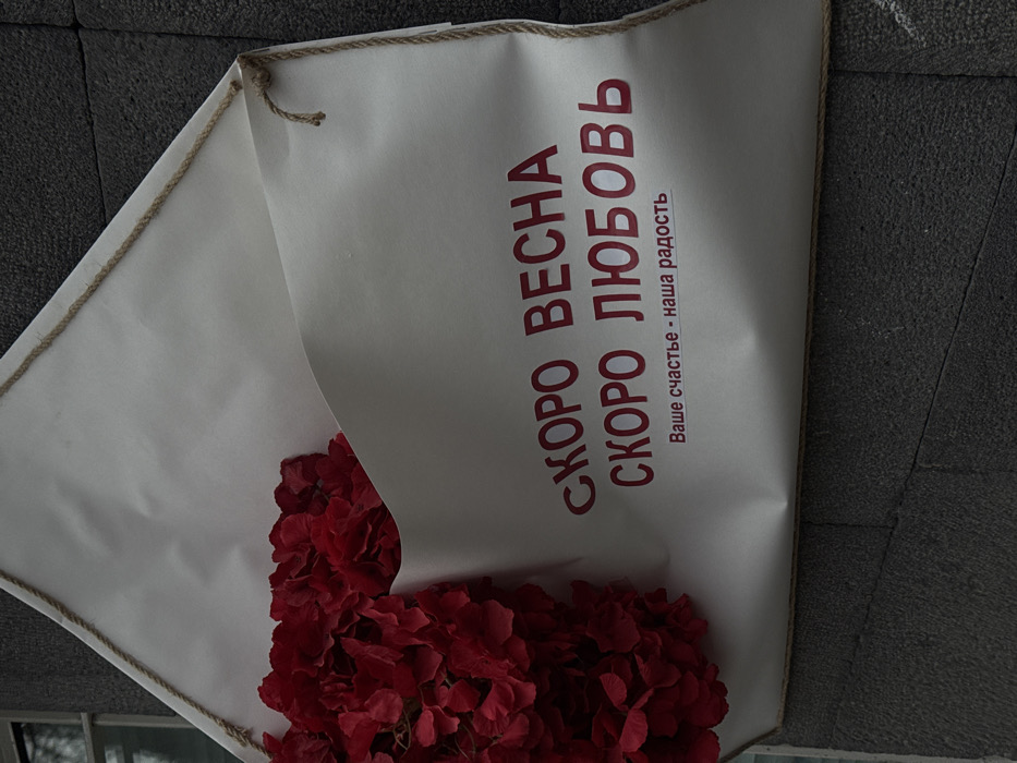 A white paper bouquet, secured with a rustic rope trim, holds a dense arrangement of red flowers. The bouquet's text reads "СКОРО ВЕСНА СКОРО ЛЮБОВЬ" (Soon Spring, Soon Love) followed by "Ваше счастье - наша радость" (Your happiness is our joy). The bouquet rests partially on a gray, textured surface, suggesting a sidewalk or stone backdrop.