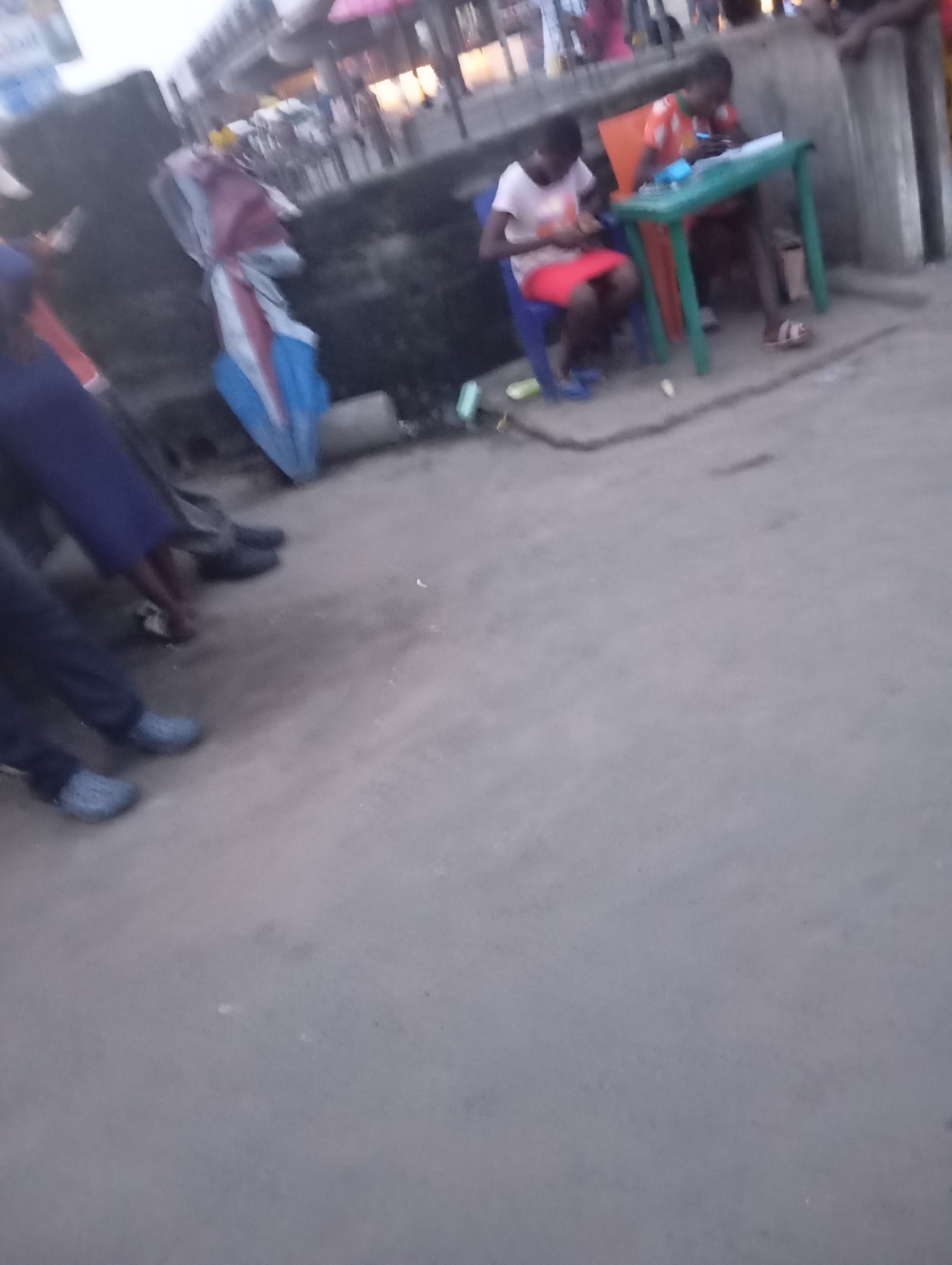 A street scene in Port Harcourt, Nigeria, shows two young people seated at a small green table. One, wearing a pink shirt and red skirt, sits on a blue chair. The other, in an orange shirt, sits on an orange chair. A blue and red bundle of cloth lies on the ground to the left. The ground is paved with concrete. In the background, a concrete wall and some blurred figures suggest an urban environment. The lighting is dim, indicating it may be early morning or late evening.