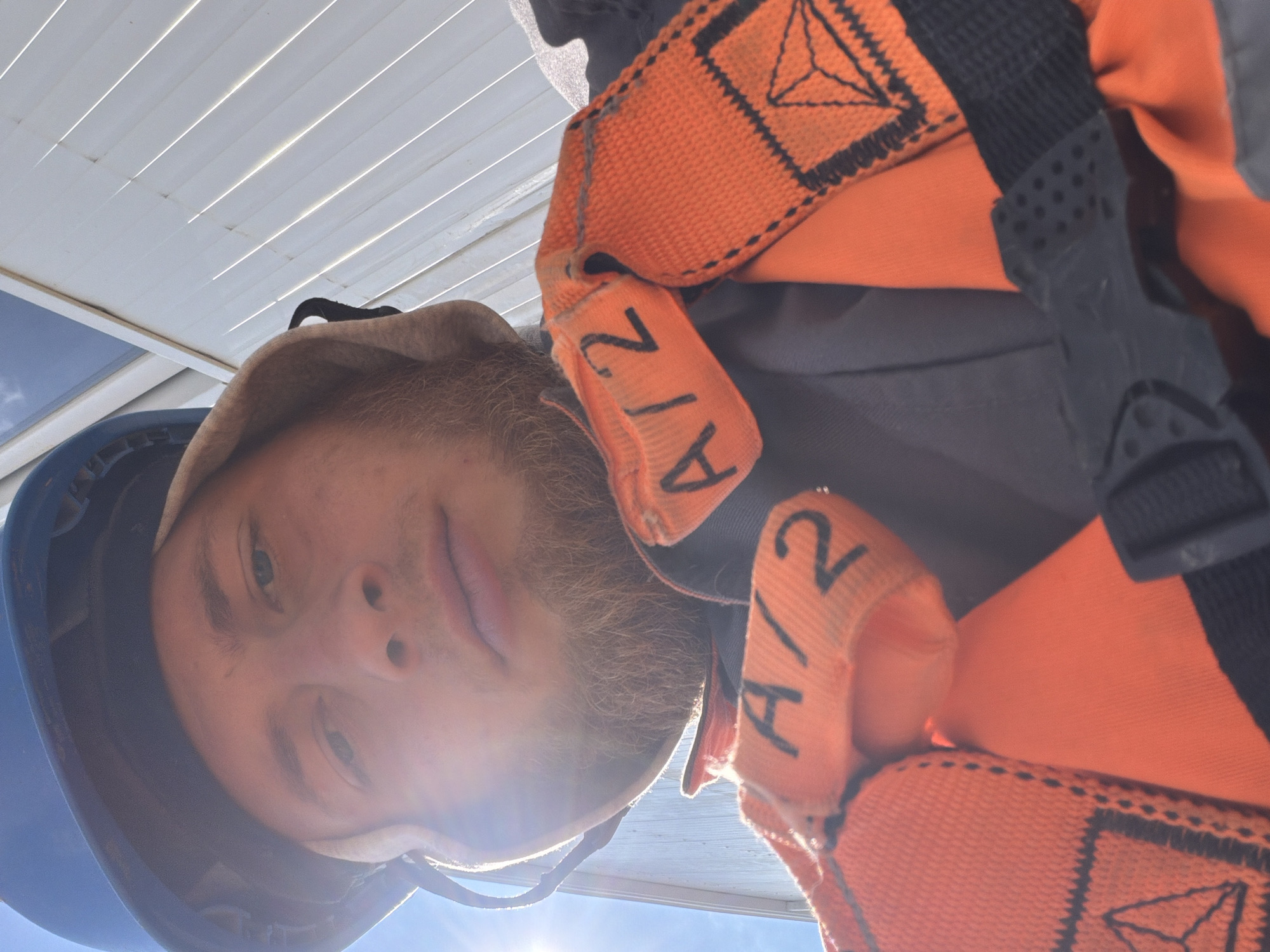 The image shows a close-up, low-angle selfie of a man wearing a blue hard hat and a tan hood. He has a beard and appears to be looking into the camera. He is also wearing an orange safety harness with black straps, which has the label "A/2" printed on it. The background consists of a white, corrugated metal structure and a bright blue sky with some clouds. The lighting suggests it is daytime, with the sun illuminating the scene from the left. The man's attire indicates he is likely on a construction site or engaged in similar work. The provided metadata indicates the location as Stróża, Poland.