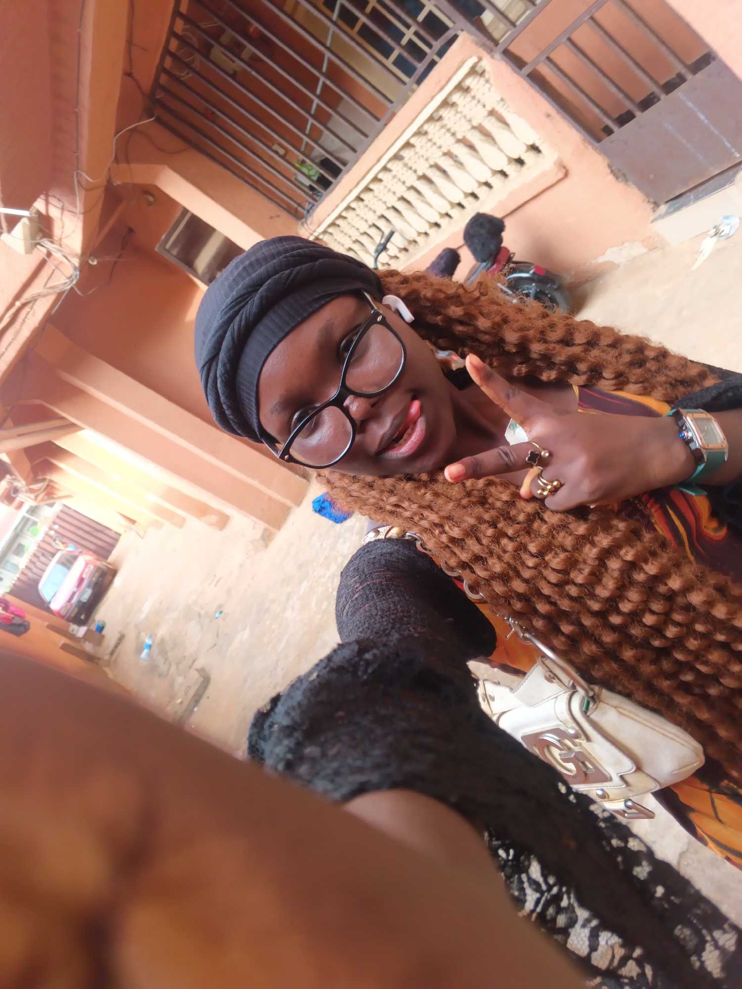 A woman with brown curly hair, wearing a black head covering, glasses, and a black lacy garment, is taking a selfie with a peace sign. She is also wearing a watch and several rings. The setting appears to be outside, possibly in a residential area, with an orange/brown building in the background, including security bars on the windows. There's a motorcycle parked behind her. The location is likely in the city of Onitsha, Nigeria.