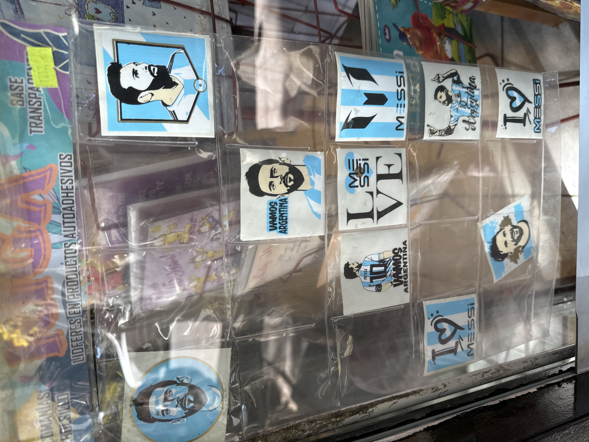 A clear photograph showcases a display of stickers in a transparent holder, likely for sale. The stickers predominately feature images related to Lionel Messi and the Argentina national football team. The stickers are in a multi-pocket display, with some pockets empty, suggesting some have been purchased or removed. The images on the stickers include a portrait of Messi with the Argentina flag, the word "Messi" in various designs, and slogans like "Vamos Argentina" (Let's Go Argentina). The display sits near a window or reflective surface, with a portion of a window frame visible. Other items, including more stationery or craft supplies, are partially visible around the sticker display. Some text is visible, including "LIDERES EN PRODUCTOS AUTOADHESIVOS" and "BASE TRANSPAR," suggesting the packaging materials. Based on the content of the stickers, the location is likely a store in Buenos Aires, Argentina.