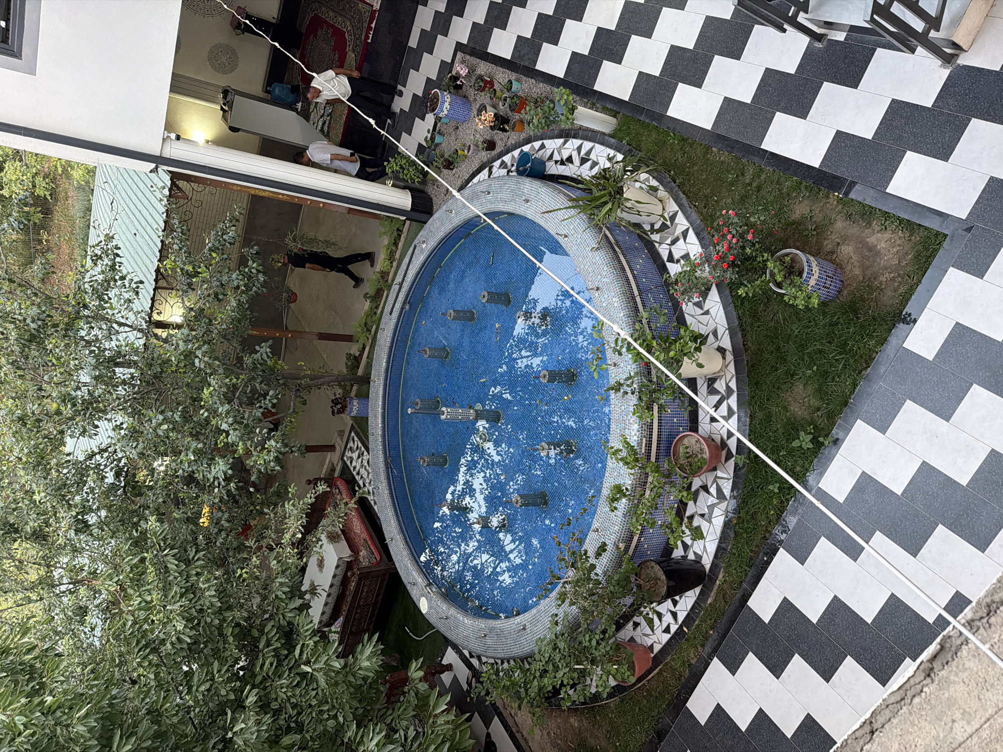 An aerial shot captures a courtyard in Nanay, Uzbekistan, featuring a circular tiled fountain with blue water and multiple spouts. Lush green trees surround the fountain, with their branches extending over the scene. A patterned tiled pathway in black and white borders the courtyard. Several potted plants, some with red flowers, are placed around the fountain and along the pathway. In the background, a building with a white exterior and a green roof is visible. Two men in casual attire are walking in the shaded area of the building, and another man is standing near a doorway. A decorative bench covered with a tablecloth is situated among the trees. The ground is a mix of grass and gravel.