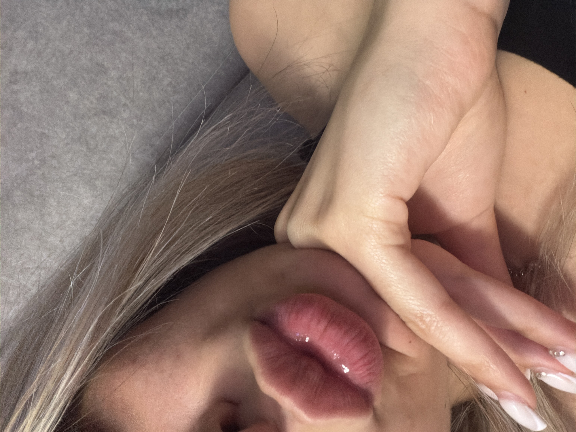The image shows a close-up of a woman's lips and chin, with her hand resting on her chin. Her lips are plump and glossy, with a pinkish-red hue. She has light-colored hair that is partially visible around her face. Her nails are manicured with a French tip and some embellishments. The background is a soft, textured gray surface. The scene appears to be indoors, with soft lighting. There is no visible information in the image to suggest a location in Ruse, Bulgaria.