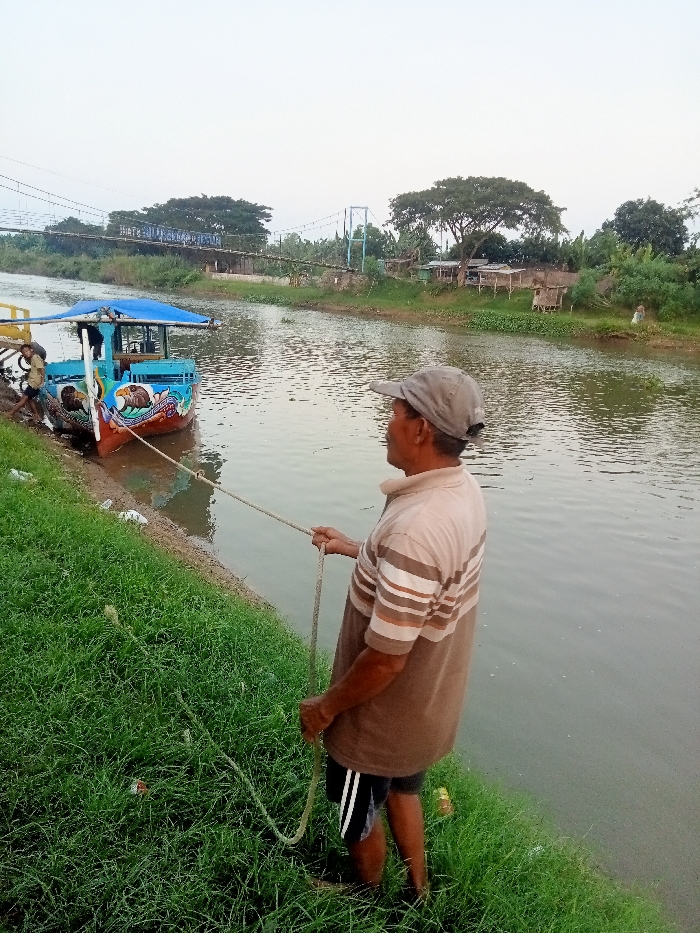 A man wearing a brown and white striped shirt and a grey cap is standing on the grassy bank of a river in Jakenan, Indonesia (latitude: -6.758, longitude: 111.075). He is holding a rope attached to a small, blue and white boat docked on the opposite bank. The boat appears to be a ferry or passenger boat, decorated with images of birds. It is located near a bridge. The river is wide and calm, with a slight current. The scene is peaceful and serene, with lush green vegetation along the riverbanks. The sky is clear and the weather is sunny.