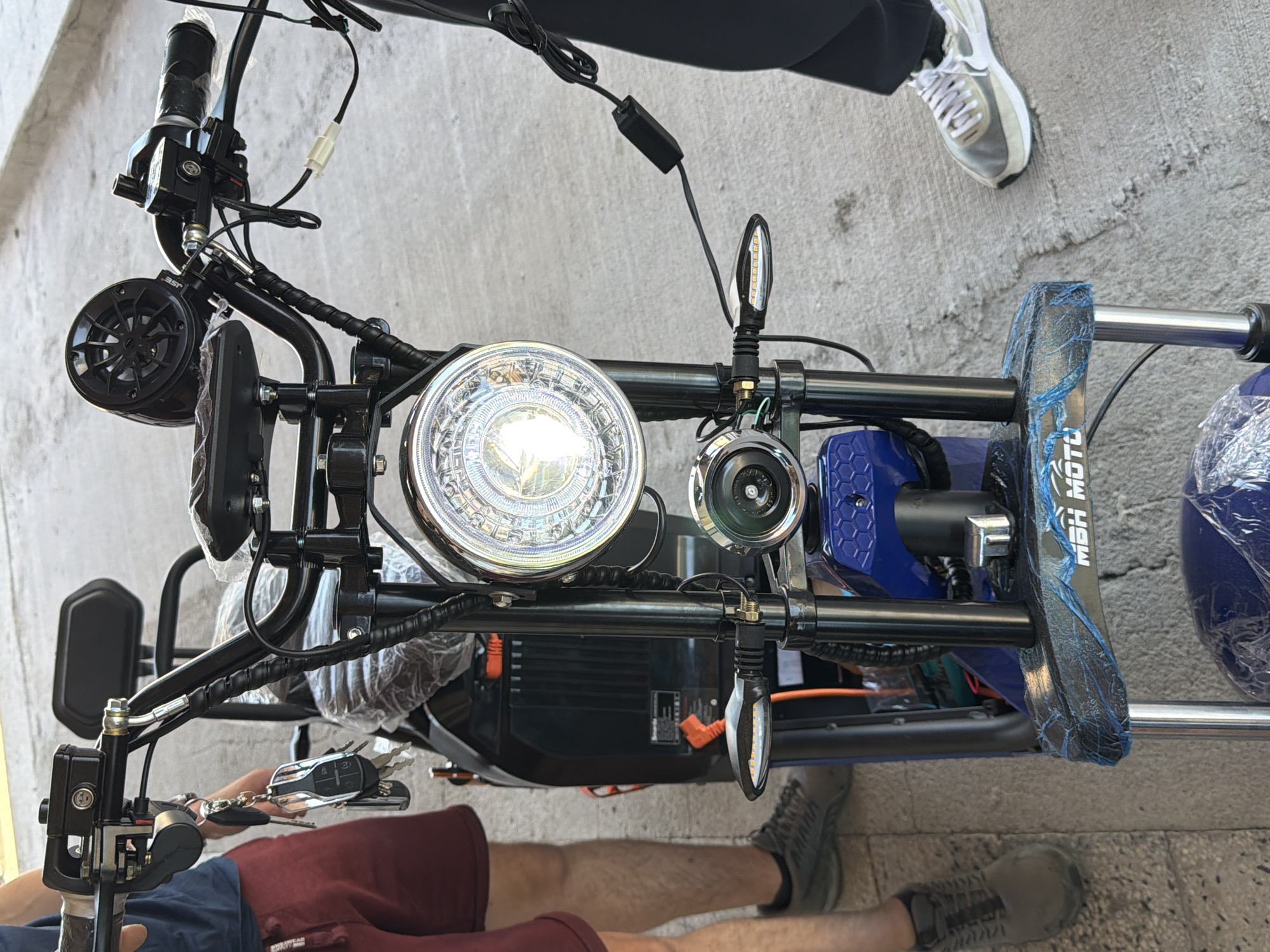 In an outdoor setting, possibly a street or sidewalk, the front view of a parked electric scooter is captured. The scooter is dark in color, with a large round headlight dominating the center of the image. It features handlebars with various controls and a small speaker mounted above. A set of keys with a remote fob hangs from the handlebars. The front of the scooter has blue accents and a protective plastic covering over a portion of the body. Two individuals are partially visible; one on the left, wearing maroon shorts and grey sneakers, and another on the right, wearing dark pants and white and grey sneakers. The ground is a rough, concrete-like surface.