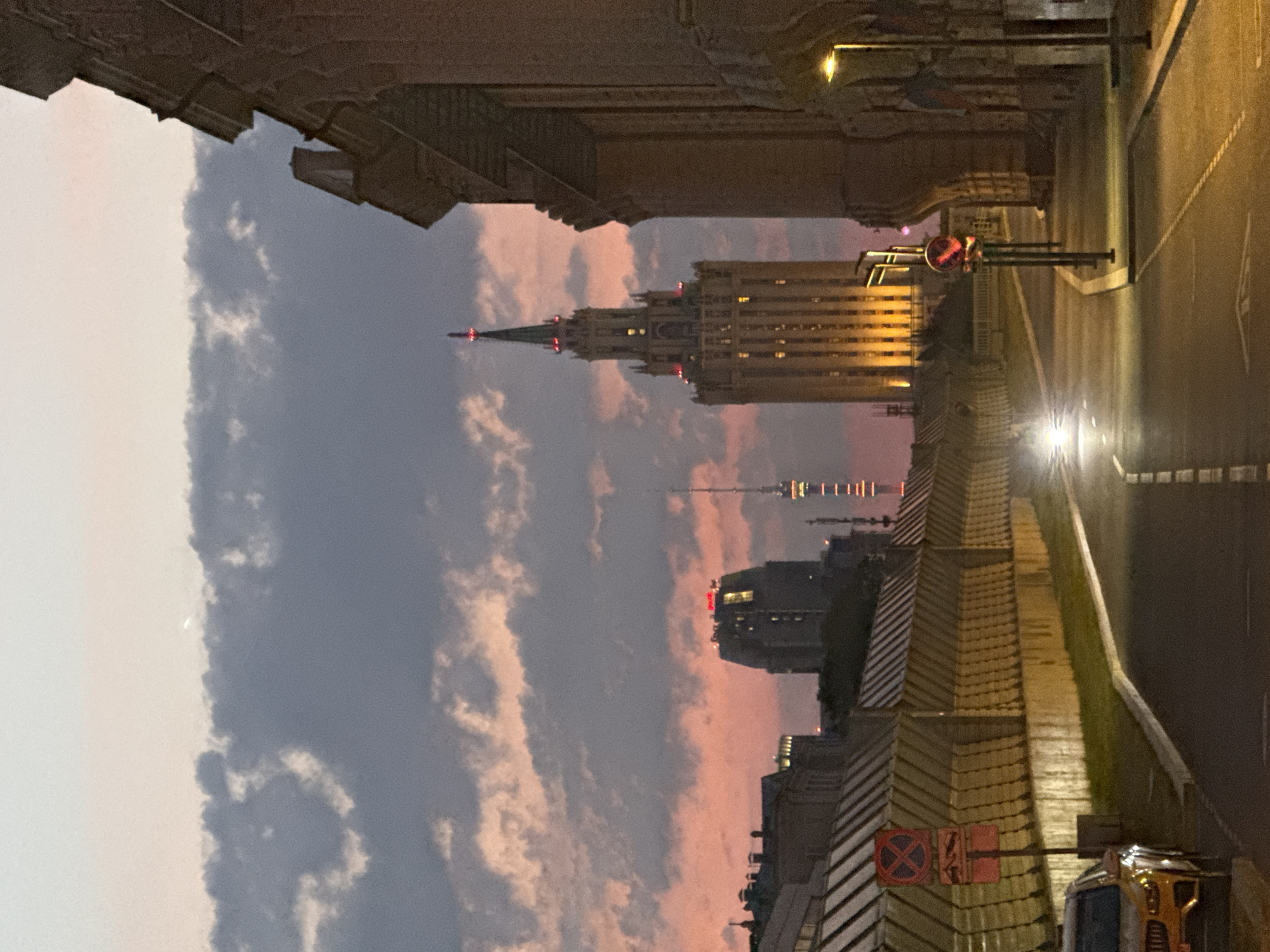 The image depicts a street scene in Moscow, Russia, at dusk. The sky is a soft gradient of pale blue and pink, with scattered grey clouds. A tall, illuminated building with a spire dominates the mid-ground, its windows glowing with warm light. To its left, another, shorter building is also lit, and in the far distance, a television tower can be seen. The foreground shows a street curving to the left, with a yellow car parked on the left side. A construction fence with yellow and grey patterns runs along the left side of the street, adorned with red traffic signs. The right side of the street is bordered by a large, stone building with arched architectural features. Streetlights are visible, casting pools of light on the pavement. The overall atmosphere is calm and urban, illuminated by artificial lights against the fading natural light.