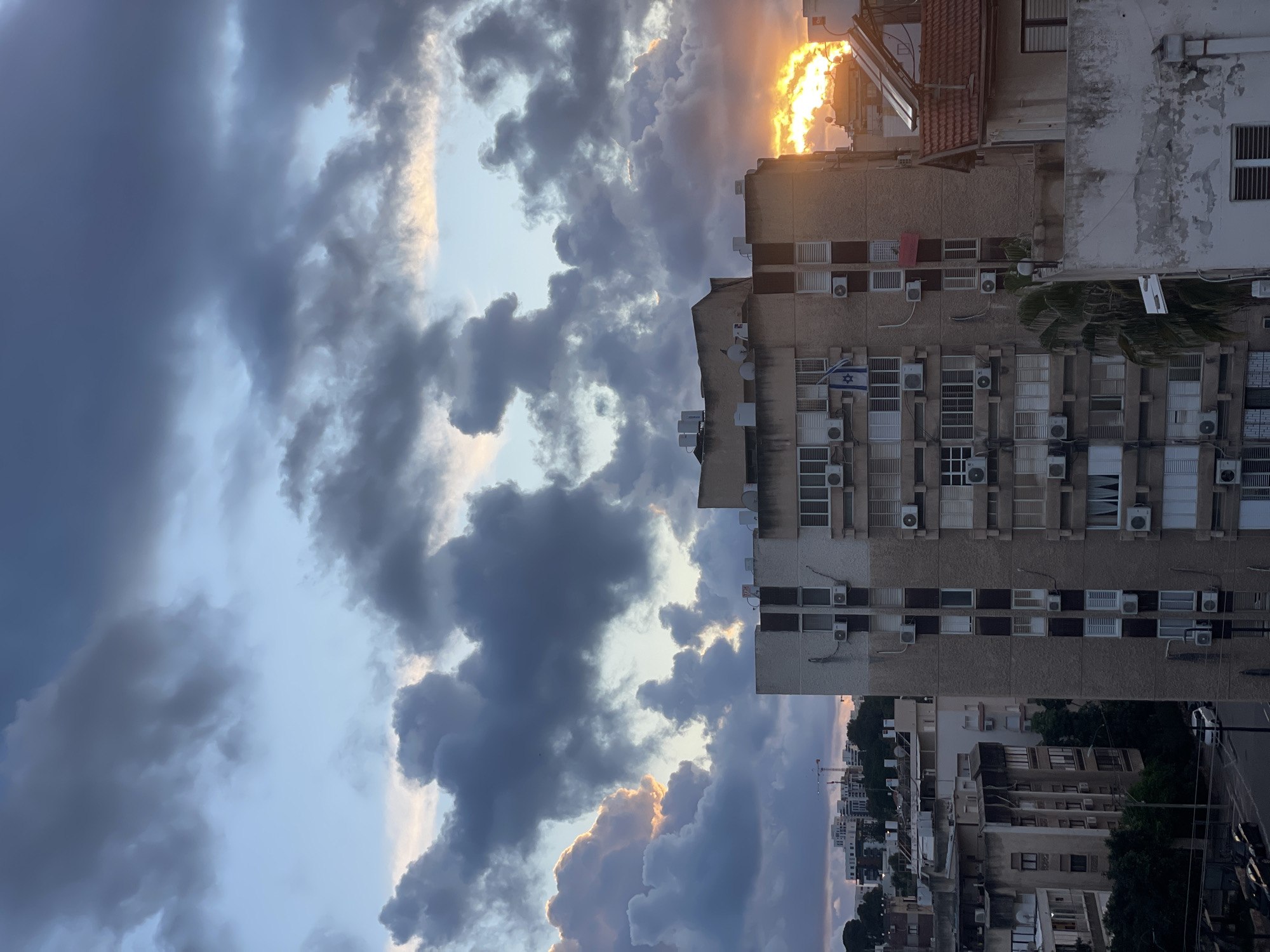 This image depicts an urban residential scene in Petah Tikva, Israel, during sunset or dusk. The sky is largely covered by dense, dark grey, and blue clouds, with the setting sun intensely visible on the right, casting a bright orange and yellow glow that illuminates cloud edges and the surrounding atmosphere.
In the midground, a prominent multi-story residential building, light beige or off-white in color with darker vertical structural elements, stands centrally. Its facade features numerous windows, many equipped with white external blinds, and a significant number of external air conditioning units are mounted below windows and on the building's surface. An Israeli flag, blue and white, is displayed vertically from a balcony or window on the left side of this building's visible face. On its flat roof, several white utility structures, likely water tanks, and satellite dishes are visible.
To the immediate right of this central building, a smaller residential structure with a sloped, reddish-brown tiled roof is visible, with a solar panel or water heater mounted on its roof, angled towards the intense light of the setting sun.
To the left of the central building, receding into the background, are several other multi-story residential buildings, varying in architectural style and condition, some appearing older and others potentially newer. Construction cranes are discernible on the distant horizon against the fading sky, indicating ongoing development. Dark green foliage, including a palm tree, is interspersed among the buildings at lower levels. A paved street runs horizontally in the lower-left portion of the image, where a white passenger vehicle, possibly a van, is visible. No individuals or specific human actions are discernible.