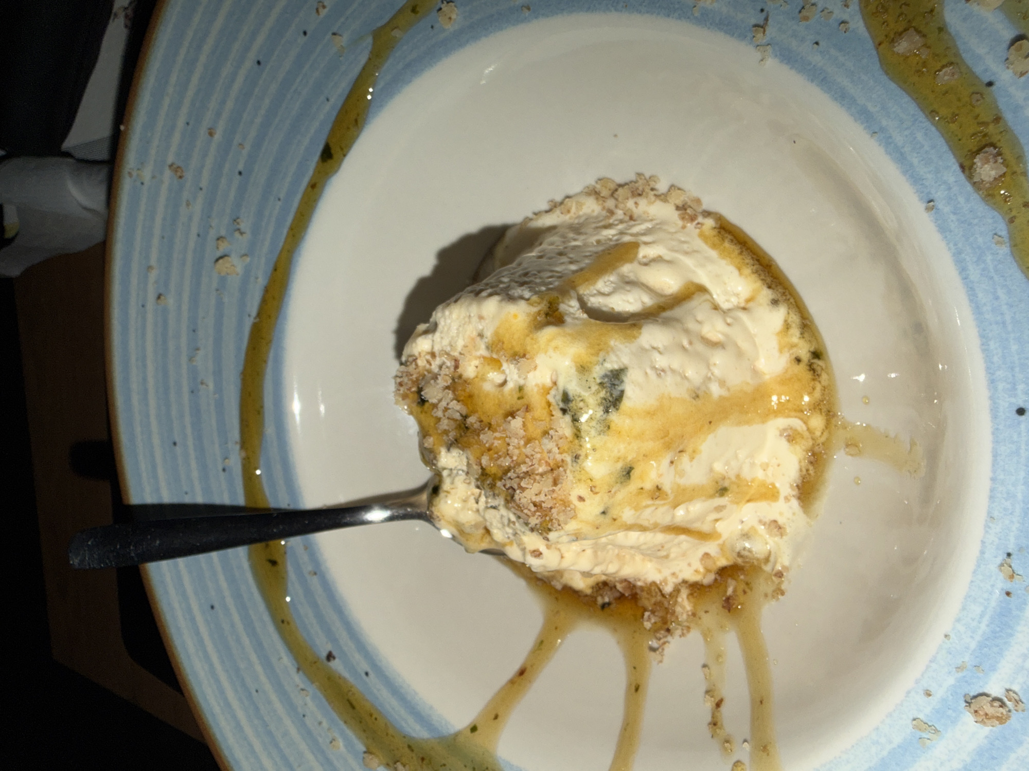 A close-up, overhead shot shows a portion of a white and blue ceramic plate containing a serving of a creamy, pale yellow dessert, possibly ice cream or cheesecake. A silver spoon is partially submerged in the dessert, and the dessert is drizzled with a golden, syrupy sauce and sprinkled with small, crunchy, breadcrumb-like pieces. The plate's rim is decorated with light blue horizontal stripes and speckled with small dark dots. The background is dark and out of focus, suggesting an indoor setting. The image is likely taken in Arbanassi, Bulgaria, given the context of the request.