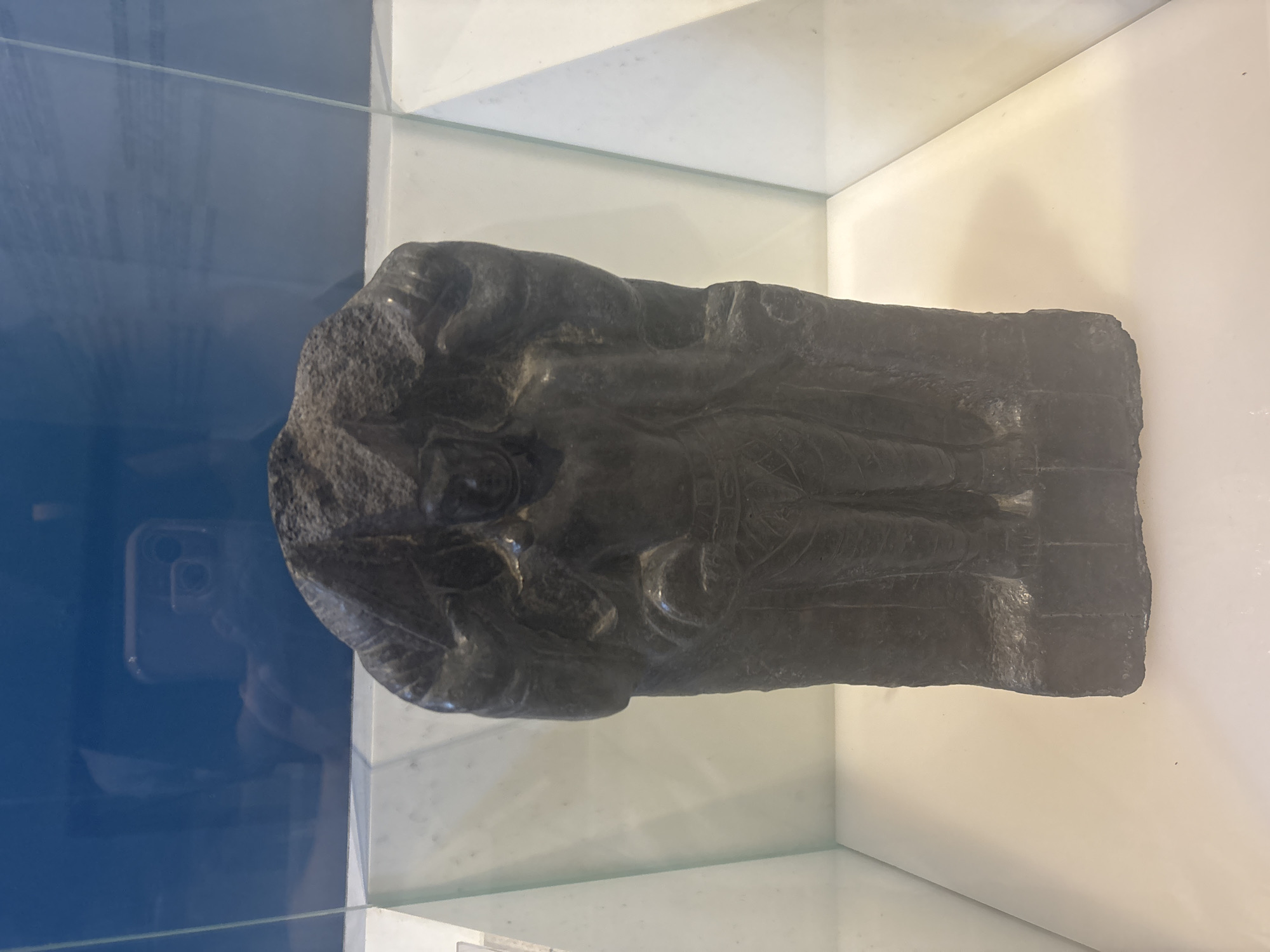 An ancient, dark stone sculpture of a standing figure is displayed in a museum exhibit. The figure is incomplete, with the head and upper body showing signs of damage or erosion. The sculpture depicts a human-like form with detailed clothing, including a waistband and patterned lower garment. The figure stands on a rectangular base. The sculpture is enclosed in a glass display case, with the reflective surface of the glass showing the museum environment, including a selfie taken by a visitor. The exhibit is located in the city of West Jakarta, Indonesia.
