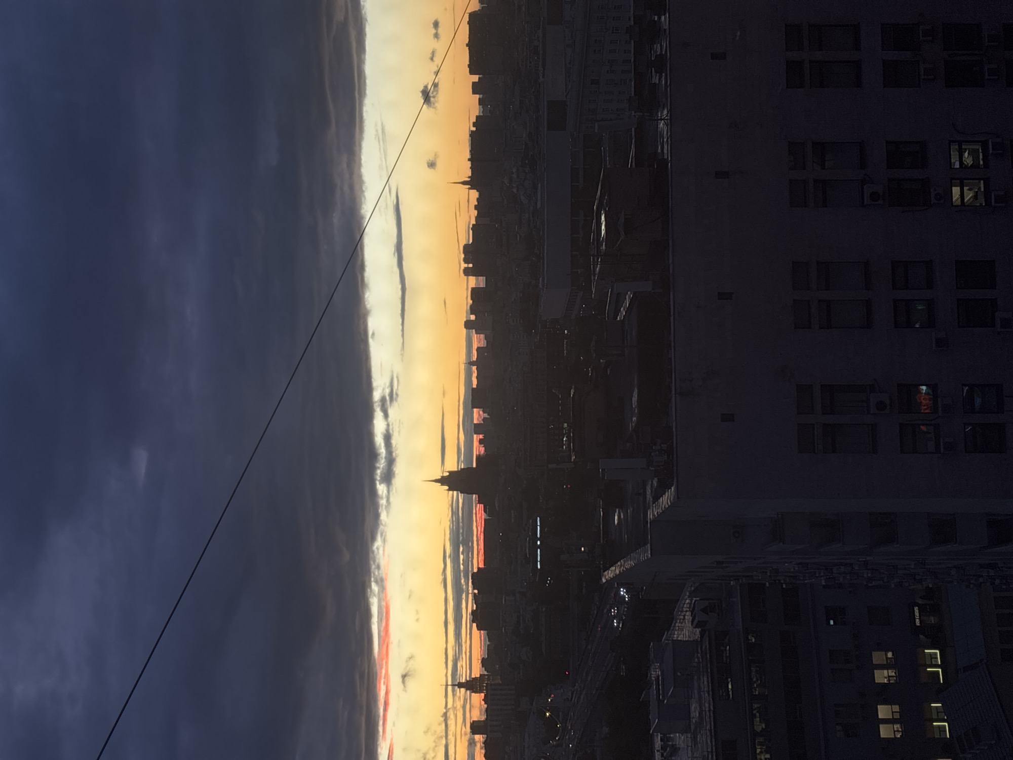 The image captures a Moscow cityscape at dusk. The sky transitions from deep blue, almost purple, to bands of orange and yellow at the horizon, suggesting a sunset or sunrise. Clouds are present in varying shades. A thin wire extends across the upper left of the frame, possibly a power line or a supporting cable. Below the sky, the cityscape is dominated by buildings of varying heights, silhouetted against the fading light. One prominent structure stands out with a spire or a tower shape. To the right, a multi-story building occupies a significant portion of the frame; its windows show some internal lighting. The lighting is dim, creating strong contrasts between light and shadow. The scene is urban and likely taken from an elevated vantage point. The presence of spired architecture suggests this could be a central area of Moscow, potentially in the vicinity of the Seven Sisters skyscrapers or other landmark buildings. Details within the skyline are difficult to distinguish due to the low light and silhouette effect.