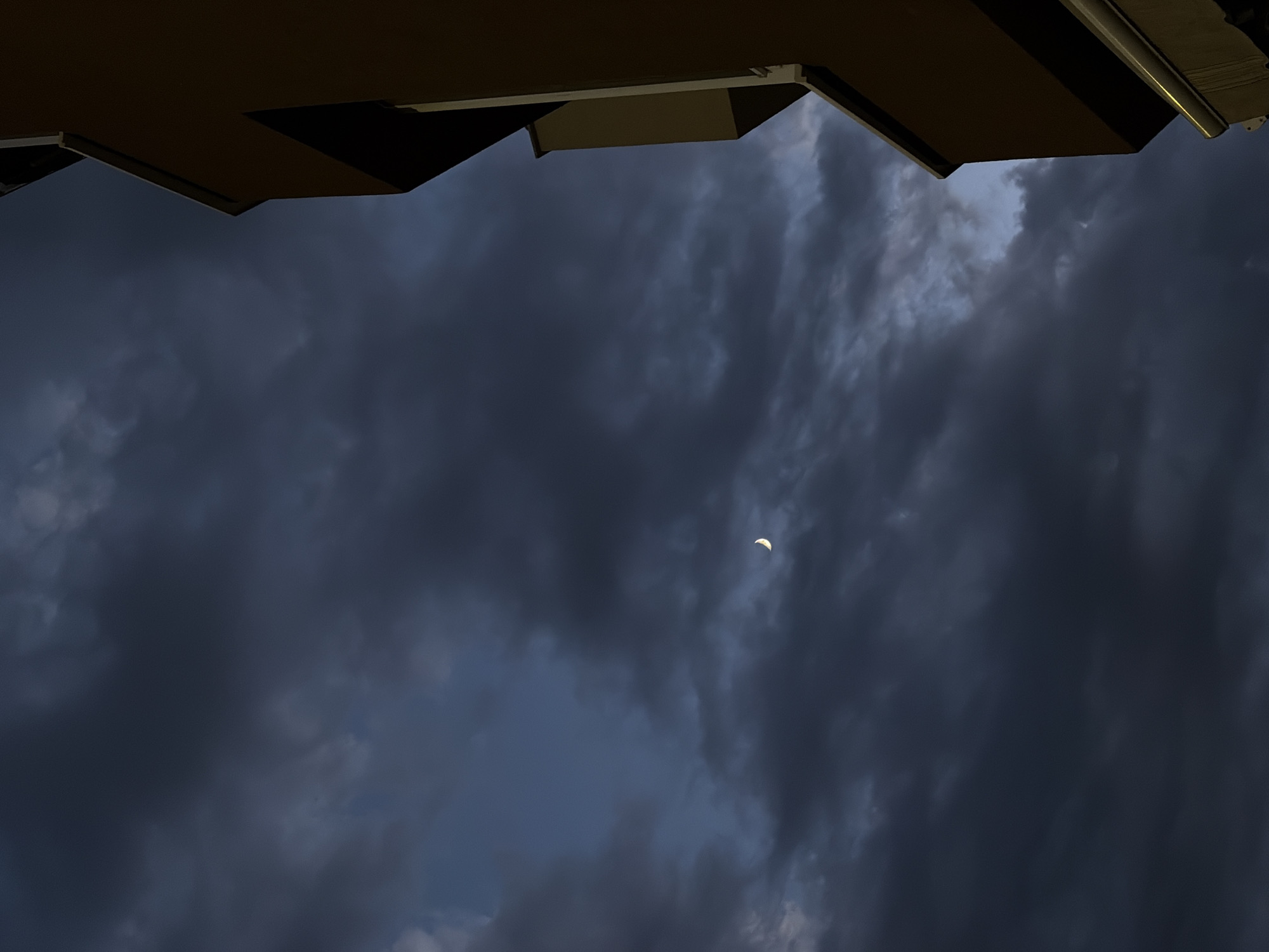 An exterior, low-angle, medium shot captures a sliver of the moon visible in a dark, cloudy sky. To the right, a portion of a modern building's facade, in shades of brown and beige, is visible, with an angled balcony or window recess. The overall mood is somber and atmospheric, suggesting twilight or early evening.