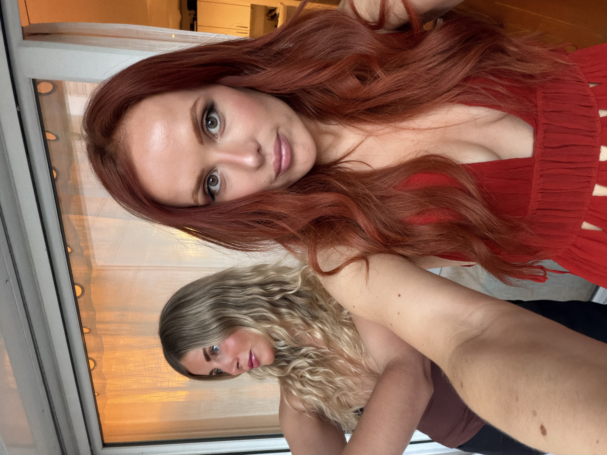 Two women pose for a selfie indoors. The woman in the foreground has long, wavy auburn hair and is wearing a red dress. Her eyes are grey. The woman behind her has long, curly blonde hair. They are positioned in front of a window. Behind them, there is a wooden floor and white cabinets. The location cannot be confirmed as Paris, France without additional information.
