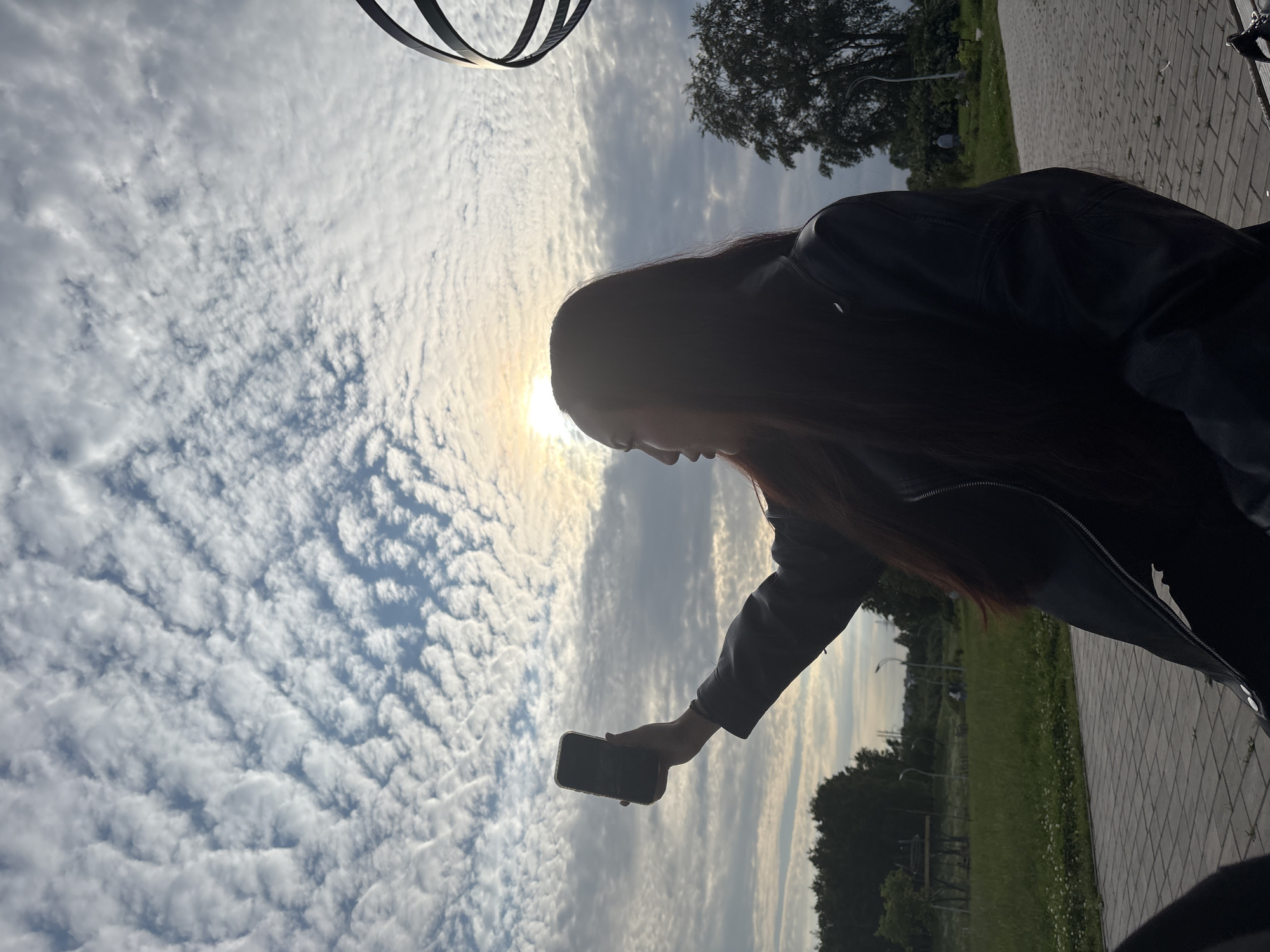 A woman in a black leather jacket is silhouetted against a cloudy sky with the sun visible behind the clouds, holding up a smartphone in her right hand as if taking a picture or video. She is facing to the left, her profile visible. She has long, dark hair. The background shows a paved walkway, a grassy area with trees, and some distant structures. The sky is filled with textured, wave-like clouds. The scene appears to be outdoors in a park-like setting.