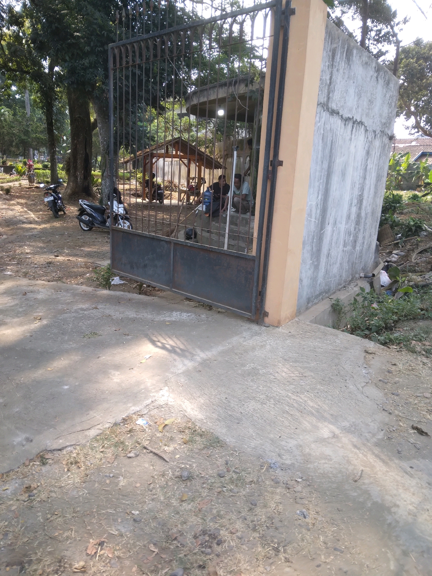 This is an outdoor, eye-level shot of a closed metal gate. To the right of the gate is a concrete wall with a beige pillar attached to the gate. Behind the gate, there is a small wooden structure with a tin roof and several people are present. Two men are crouched down near the gate, one in a light blue shirt and the other in a black shirt. Another man in a black shirt is standing behind them. Two motorbikes are parked to the left of the gate, and trees and vegetation are visible in the background. The ground in the foreground is a mix of concrete and dry soil with scattered leaves and small stones. The scene appears to be in a park or public area within Desa Pamijen Kecamatan Sokaraja, Indonesia, given the lighting and vegetation.