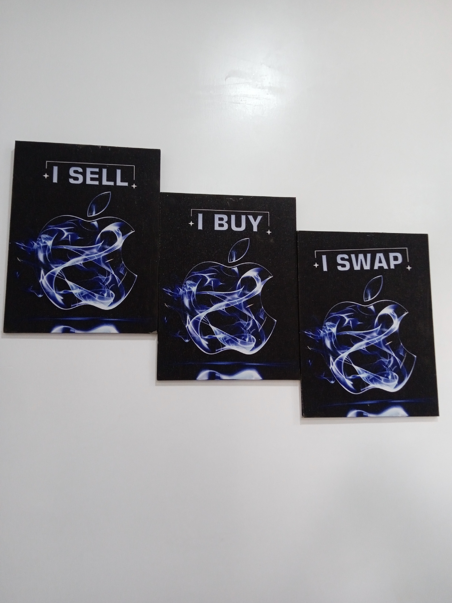Three black cards with the Apple logo are arranged on a white surface. Each card features the logo in a swirling blue design and the phrases "I SELL", "I BUY", and "I SWAP" respectively. The cards are placed side by side in that order. No people are visible. The location is not identifiable as Jos, Nigeria.
