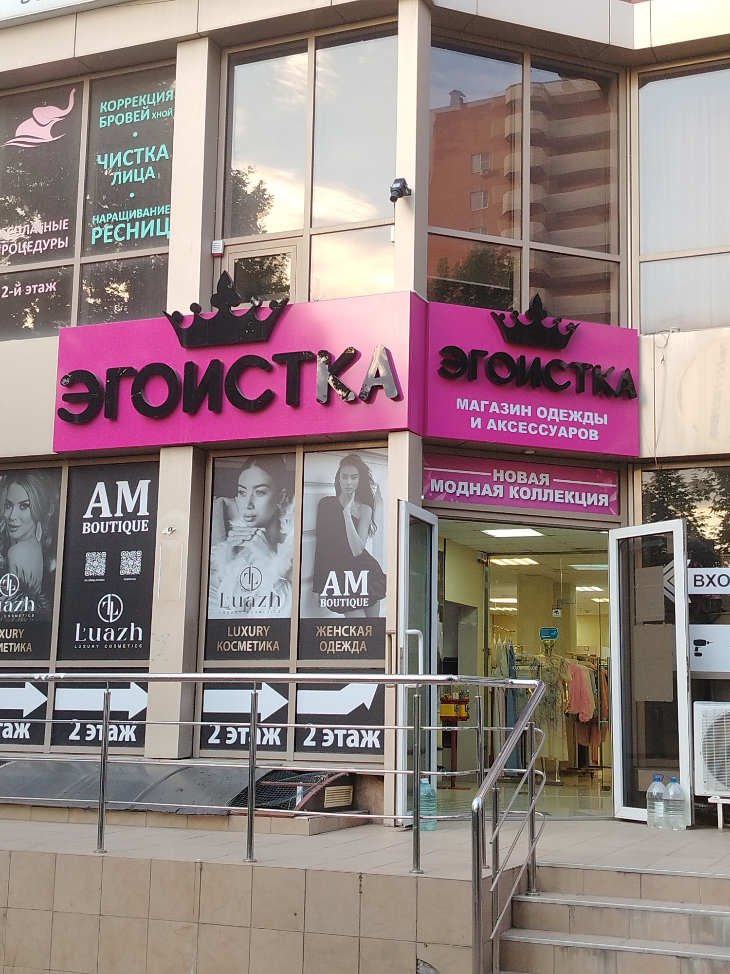 A storefront in Krasnodar, Russia, features a large pink sign with black lettering that reads "ЭГОИСТКА" (Egoistka) in stylized font, topped with a black crown graphic. Below this, smaller text indicates "МАГАЗИН ОДЕЖДЫ И АКСЕССУАРОВ" (Clothing and Accessories Store). Further down, a banner announces "НОВАЯ МОДНАЯ КОЛЛЕКЦИЯ" (New Fashion Collection). The entrance to the store is open, revealing mannequins displaying clothing inside. To the left of the main sign, another window display shows advertisements for "AM BOUTIQUE" and "Luazh LUXURY COSMETICS," with QR codes and images of women. Above these displays, text on the window reads "КОРРЕКЦИЯ БРОВЕЙ хной" (Eyebrow correction with henna), "ЧИСТКА ЛИЦА" (Facial cleansing), and "НАРАЩИВАНИЕ РЕСНИЦ" (Eyelash extensions). Arrows pointing to the right are visible on the lower portion of the storefront, with text indicating "2 ЭТАЖ" (2nd floor). The building exterior is a mix of light-colored panels and large glass windows, with a security camera visible on the left side. Several large bottles of water are placed near the entrance.