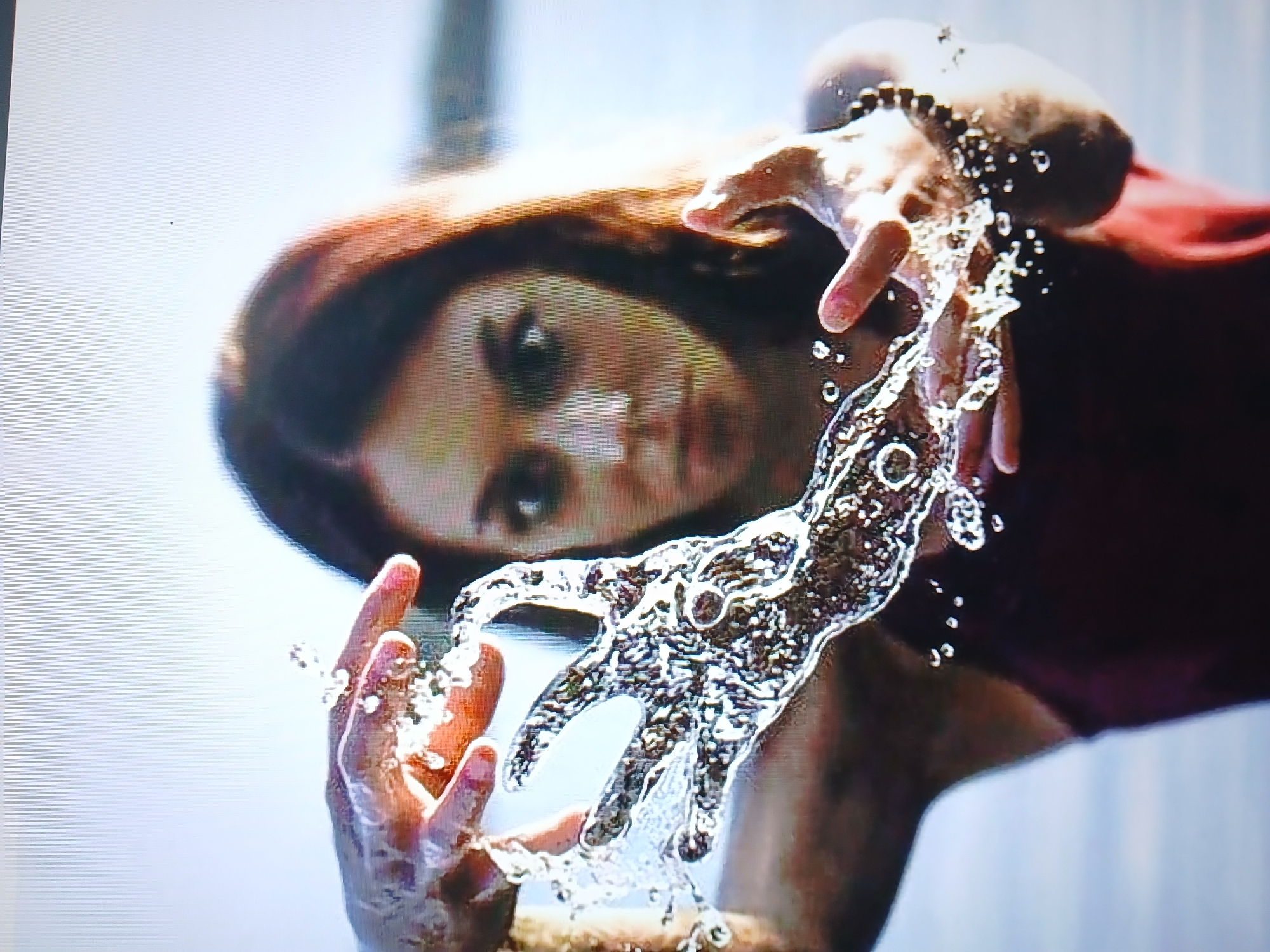 A medium shot captures a young woman with reddish-brown hair and wide eyes, holding her hands in front of her face. Water is being manipulated between her hands, forming a shape that resembles an open palm with elongated fingers. Her right hand is blurred by the water, while her left hand is more clearly visible, partially submerged in the water. She is wearing a dark red sleeveless top. The background is out of focus, showing a bright sky and what appears to be a body of water. The scene is well-lit, suggesting it is outdoors during the day.