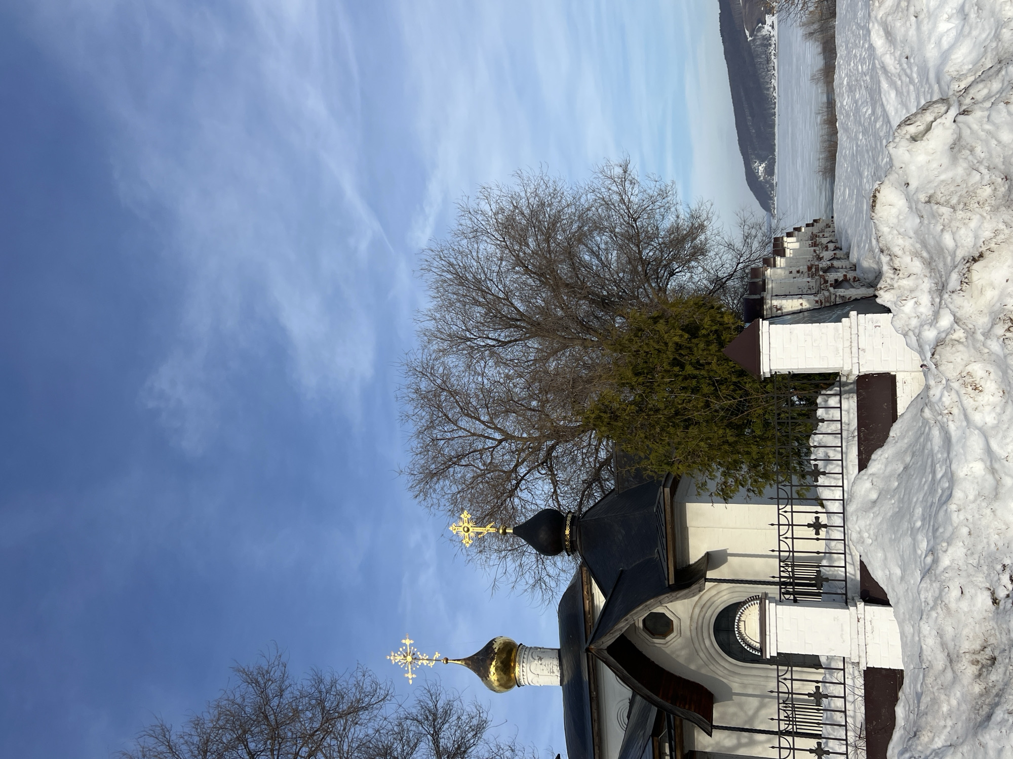 This image depicts a winter landscape in Sviyazhsk, Russia. In the immediate foreground, large, irregular mounds of dirty snow are visible. Behind these snowdrifts, a white brick fence with dark, pyramid-capped pillars and black wrought-iron sections, each featuring a cross motif, runs horizontally across the midground.
Behind the fence, parts of a white-walled religious edifice, likely a church or monastery, are visible. This structure features a dark roof and an arched entrance or window detailed with dark trim, along with a small octagonal window. Two distinct cupolas rise above the building: one larger, golden onion dome topped with a golden Orthodox cross, and to its right, a smaller, black dome also surmounted by a golden Orthodox cross. Bare deciduous trees with intricate branch patterns are scattered throughout the scene, primarily on the left and behind the buildings, while a dark green evergreen tree stands adjacent to the smaller dome.
Beyond the architectural elements and trees, a vast expanse of frozen water, appearing as a flat, snow-dusted surface, stretches to the horizon. In the far background, low, snow-dusted hills or banks, sparsely covered with dark vegetation, line the distant edge of the frozen body of water. The sky above is a clear, bright blue with scattered wispy white clouds and visible contrails. No individuals are present in the scene.