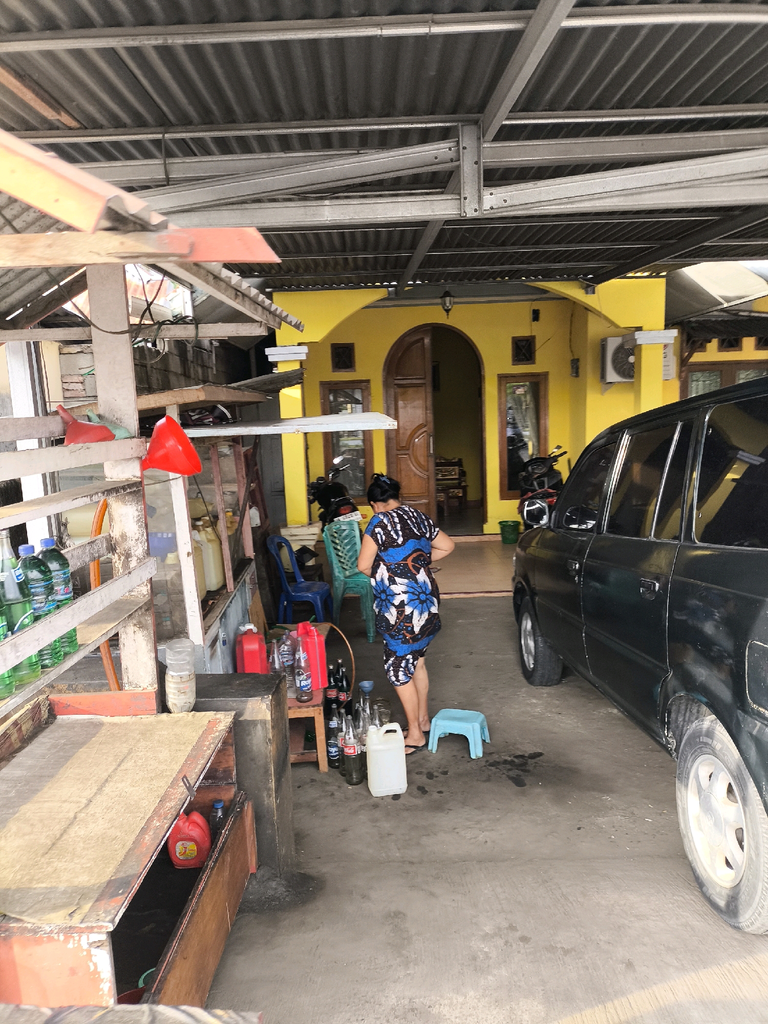 The image depicts an outdoor scene in Gorontalo, Indonesia, possibly a roadside stall or a small shop. A woman wearing a colorful patterned dress is standing in the center of the frame, facing away from the camera and towards a yellow building. She appears to be engaged in some activity related to the items spread around her.
In the foreground, to the left, there are several shelves stacked with bottles of green liquid, likely beverages. Next to these shelves, a red funnel is prominently displayed. Further to the right, there are various containers, including red jerry cans, plastic bottles, and what appear to be large white liquid containers. A dark-colored car is parked on the right side of the frame, partially obscuring the view of the yellow building.
The yellow building in the background features an arched doorway and windows, with an air conditioning unit mounted on the exterior wall. Motorcycles are visible parked near the building. The overall environment suggests a warm climate, with the corrugated metal roof structure indicating a functional, perhaps makeshift, shelter. The ground is paved with concrete, and there are some dark stains on the surface, hinting at spilled liquids. The scene appears to be a commercial setting, possibly selling drinks or small goods, with the car suggesting a point of transaction or a customer.