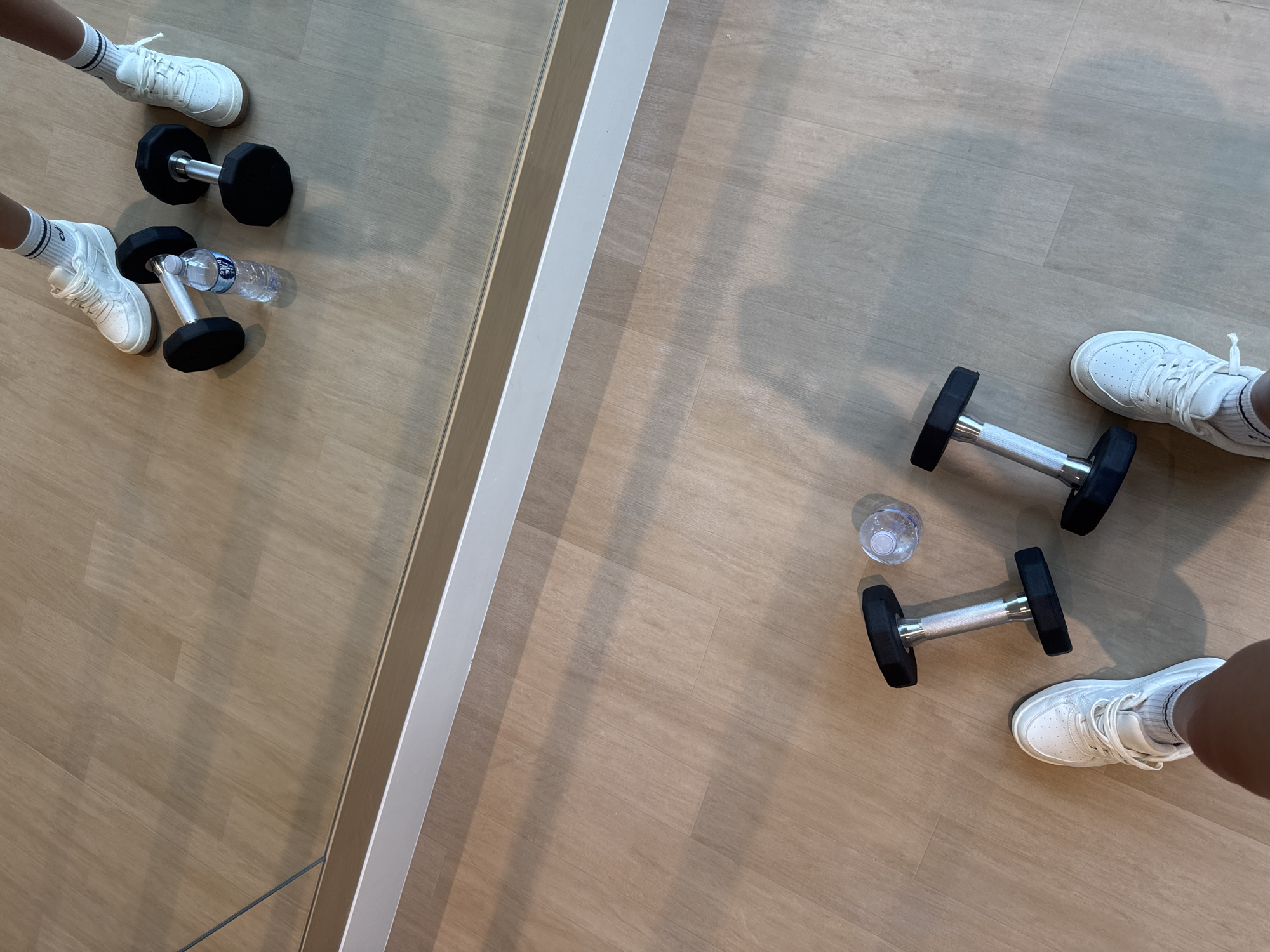 In a gym, a pair of white sneakers and two dumbbells are arranged on a wooden floor. A bottle of water is placed between the dumbbells. In the reflection of a mirror, another pair of white sneakers and dumbbells are visible. The scene appears to be staged for a workout session.