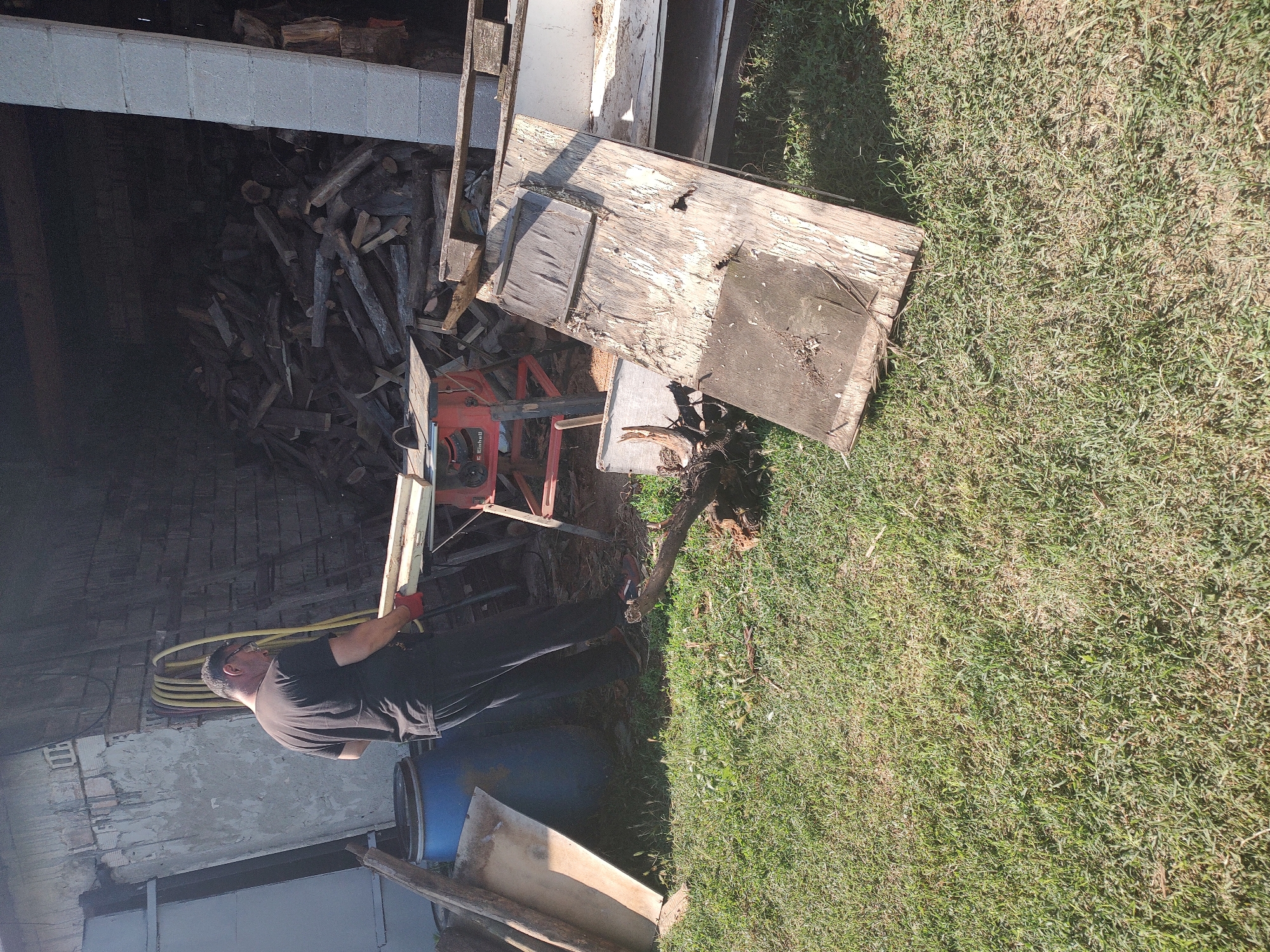 The image depicts an outdoor scene in Kochmar, Bulgaria. A middle-aged man wearing a black t-shirt and black pants is standing next to a red table saw. He is wearing red gloves and holding two wooden planks. Behind him, a large pile of firewood is visible inside an open-fronted structure. To the left of the man, a blue barrel and several pieces of wood are present. The ground is covered in green grass. The scene appears to be a work area or yard.
