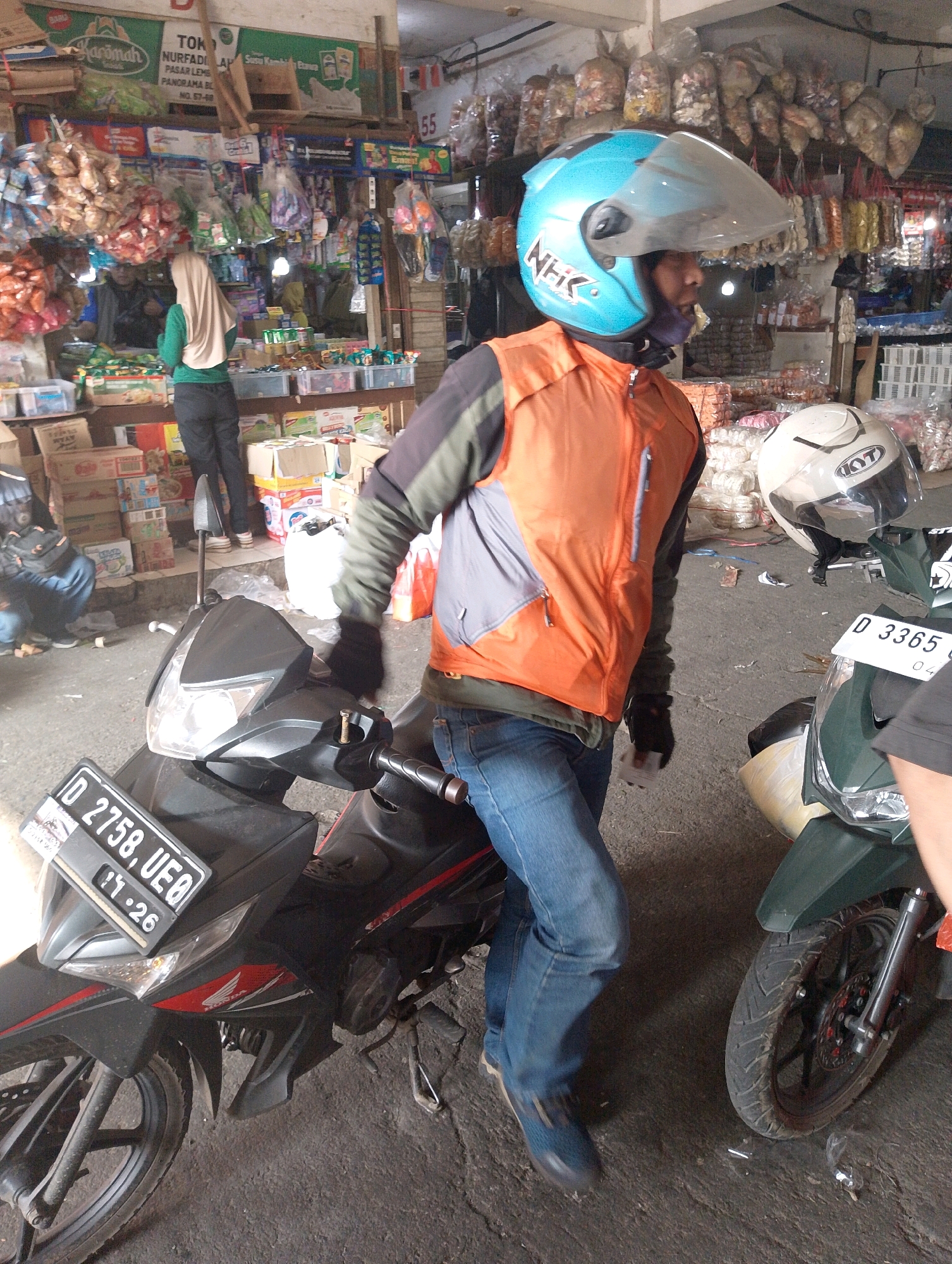 The image depicts a street market scene in Lembang, Indonesia. In the foreground, two motorcycles are visible. The motorcycle on the left is black with red accents and has a license plate reading "D 2758 UEQ". The motorcycle on the right is green and has a license plate reading "D 3365". A person wearing a blue helmet with a clear visor, an orange vest over a dark jacket, and blue jeans is standing next to the black motorcycle. They are wearing black gloves. Another person, mostly obscured, is standing next to the green motorcycle. In the background, stalls are filled with various goods, including packaged snacks and dried foods hanging from the ceiling. Several boxes and crates are stacked behind the stalls. A woman wearing a beige hijab and a green shirt is visible in the background, interacting with the market. The ground is paved with asphalt. The overall environment suggests a busy, open-air marketplace.