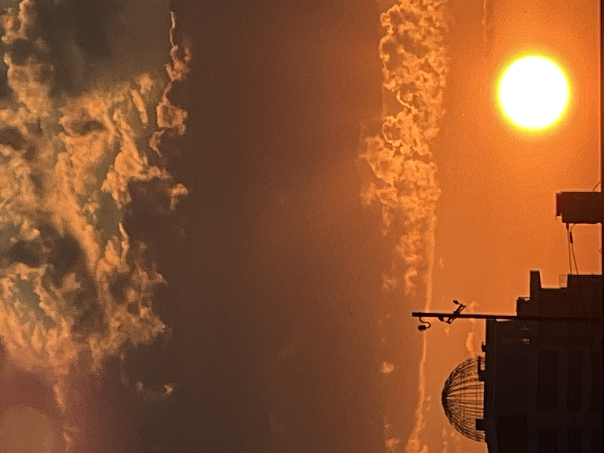 This is a nighttime, outdoor, low-angle shot of a cityscape at sunset. The sun is a bright, luminous orb in the upper right quadrant of the frame, casting a warm, orange glow over the scene. Dramatic, wispy clouds are visible in the sky, illuminated by the setting sun. In the foreground, silhouetted against the sunset, is a building with a distinctive domed structure on top. The building's dark silhouette contrasts sharply with the vibrant sky, creating a striking visual. The overall mood is serene and majestic, capturing the beauty of a sunset over an urban landscape. There is no visible text in the image. The location is inferred to be Krasnodar, Russia.