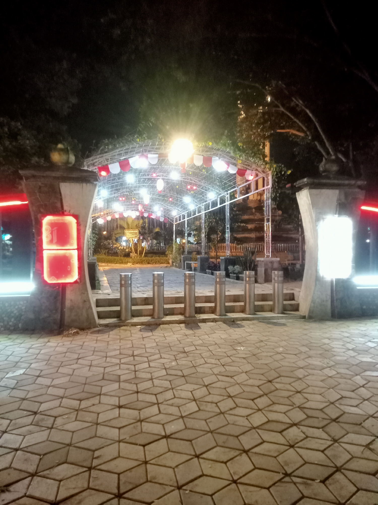 The image depicts an outdoor scene at night in Tulungagung, Indonesia. The foreground features a paved walkway with a geometric pattern of interlocking hexagonal tiles. A pathway of grey concrete steps leads up to a covered archway. Along the steps, several cylindrical metal bollards are arranged in a row.
The archway is illuminated by a string of red and white pennant lights and numerous small white bulbs, creating a festive atmosphere. The structure of the archway appears to be made of metal. Flanking the entrance to the archway are two large, stone-like pillars. The pillar on the left has a prominent square red illuminated sign with the number "31" displayed in illuminated white characters. The pillar on the right has a bright white illuminated panel. Trees with dark foliage are visible behind the archway, and some appear to be a part of the illumination. In the background, there are more lights and structures suggesting a public area, possibly a park or event space. The overall impression is of a well-lit, decorated area at night in Tulungagung.