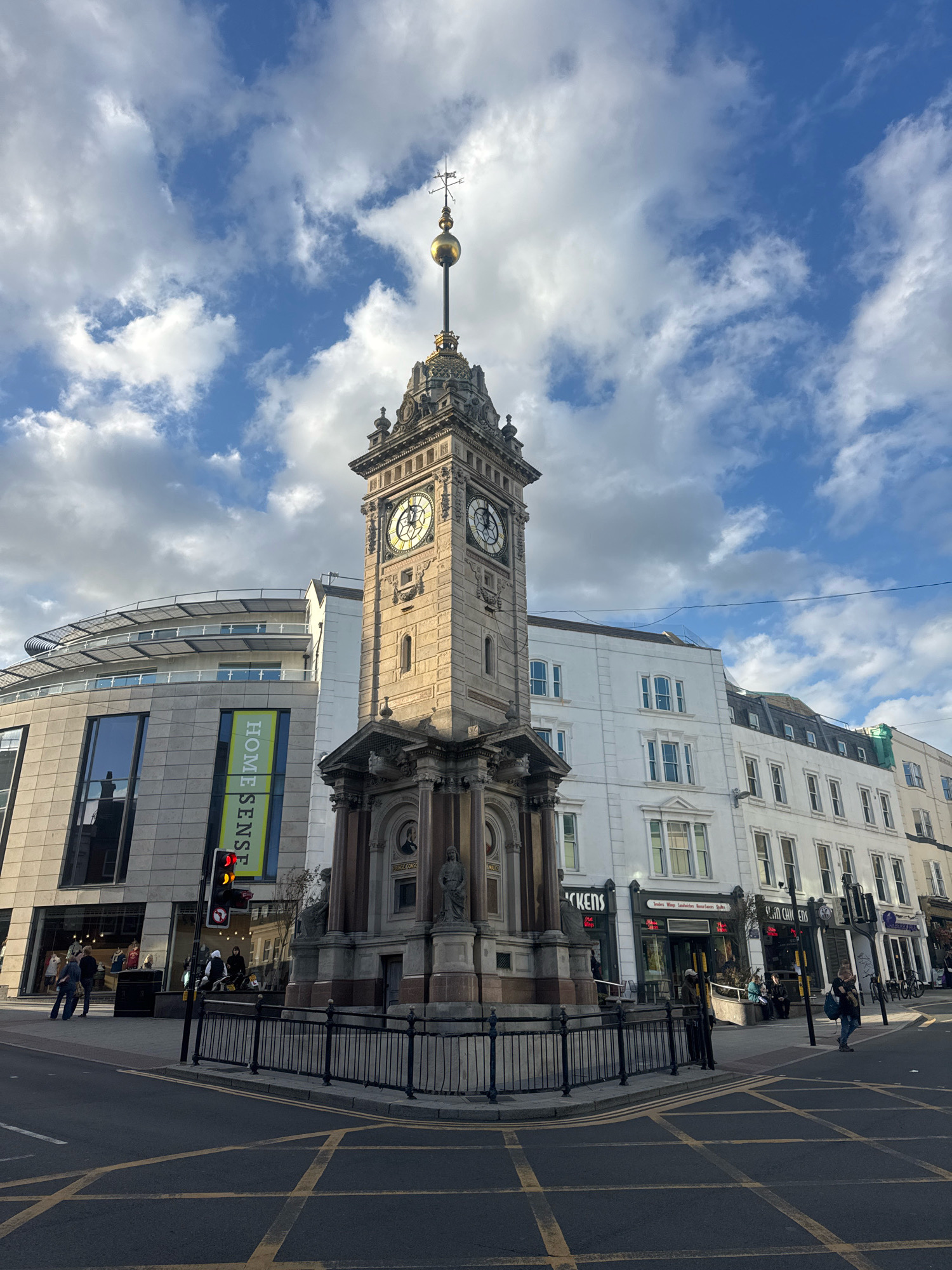 A wide-angle, eye-level shot captures the Royal Pavilion Gardens Clock Tower in Brighton, United Kingdom, bathed in the warm light of a late afternoon sun. The ornate, stone clock tower, adorned with multiple clocks and architectural details, stands prominently in the foreground, dominating the center of the frame. A golden orb and weather vane crown its peak.
To the left of the clock tower, a modern, curved building with large glass windows houses a "HOME SENSE" store, its banner prominently displayed. A traffic light with red and yellow signals indicates a pedestrian crossing. Beyond the clock tower, a row of traditional white buildings with multiple windows lines the street, interspersed with shops.
The sky above is a vibrant blue, punctuated by fluffy white clouds, suggesting fair weather. The street in the foreground is paved with asphalt and marked with yellow criss-cross lines, indicating a no-stopping zone. Several people are visible throughout the scene, walking on the sidewalks, browsing in shops, and waiting at the traffic light. A black metal fence encloses the base of the clock tower. The scene depicts a bustling urban environment on a clear day.