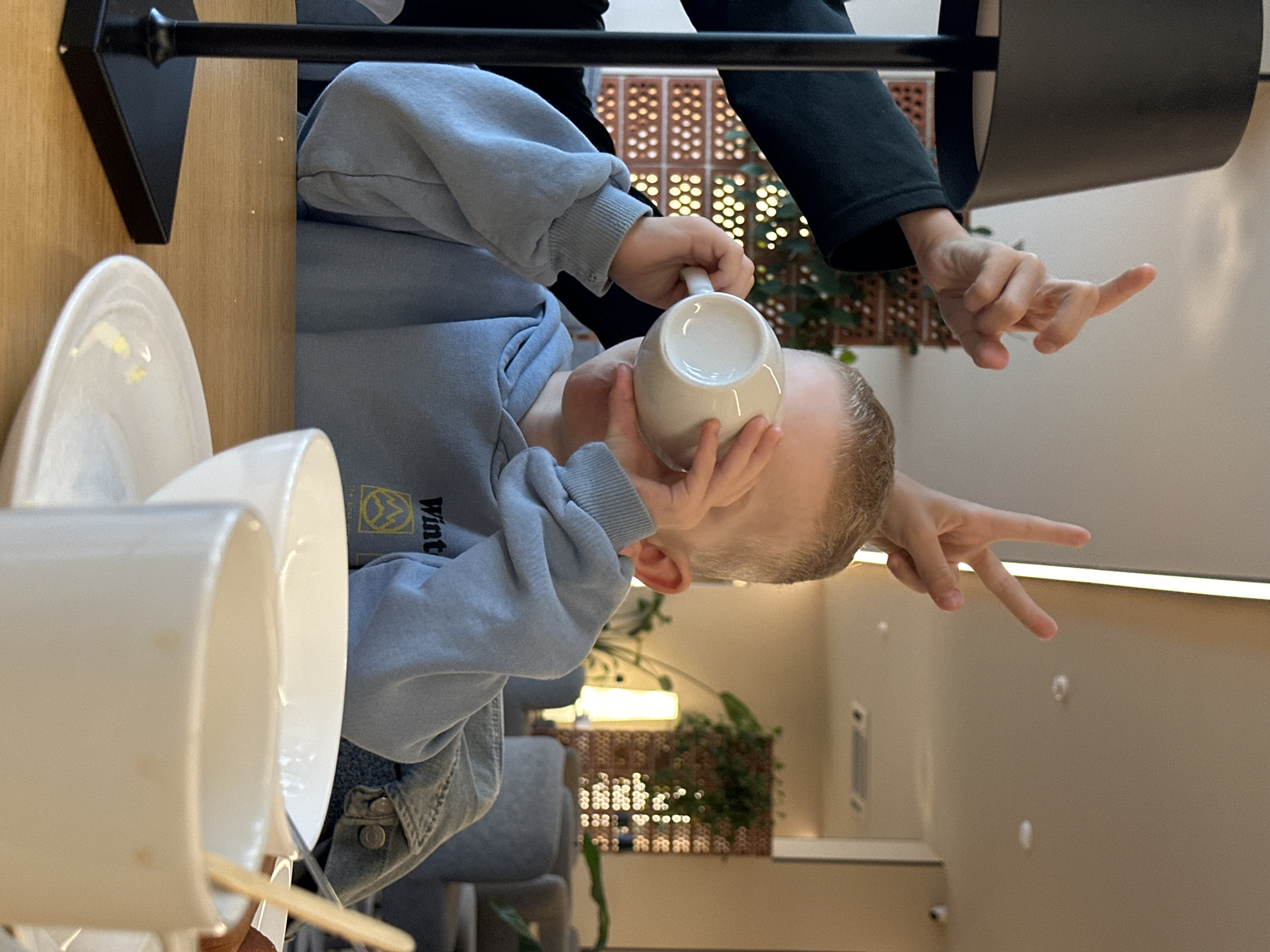 In Bolshoye Savino, Russia, a young boy is seated at a table with a light blue hoodie and a denim jacket, drinking from a white mug. Two hands are visible above him, one making a "bunny ears" gesture with two fingers, and the other pointing upwards. On the table, there is a white saucer, a large white bowl, and another white mug with a wooden stirrer. A black lamp base is on the left side of the frame. The background suggests an indoor setting, possibly a cafe or restaurant, with blurred furniture and plants.