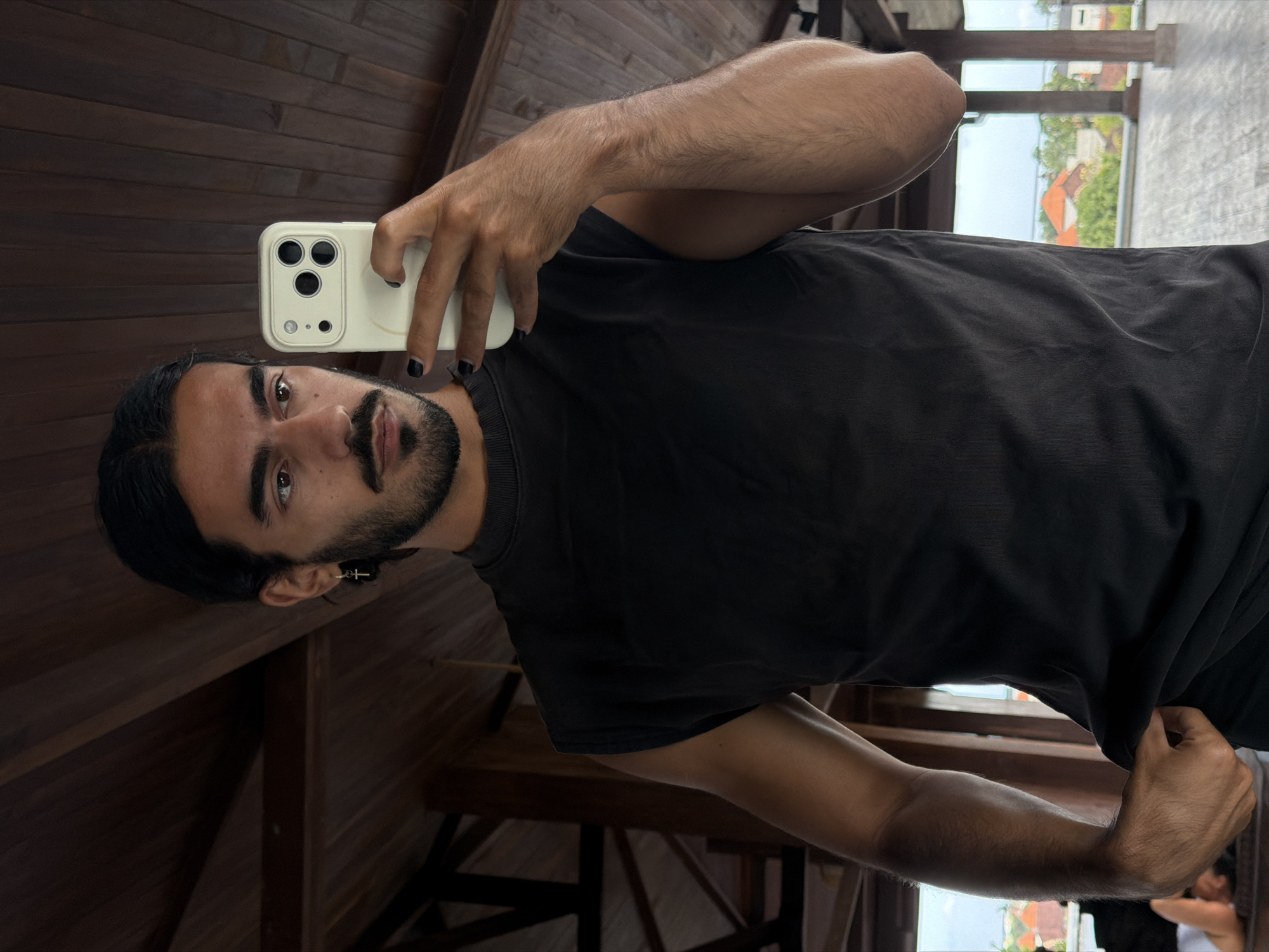 A young man with dark hair and a beard takes a selfie in a room with a wooden ceiling. He is wearing a black sleeveless shirt and holding a cream-colored phone in his right hand. The room appears to be on an upper floor, as a blurred view of rooftops and greenery is visible through windows in the background. The architectural style suggests a tropical location, possibly in or around Mengwi, Indonesia. The man's left hand is tucked into the waistband of his pants. He is wearing a small cross earring in his left ear and has black nail polish on his fingernails.