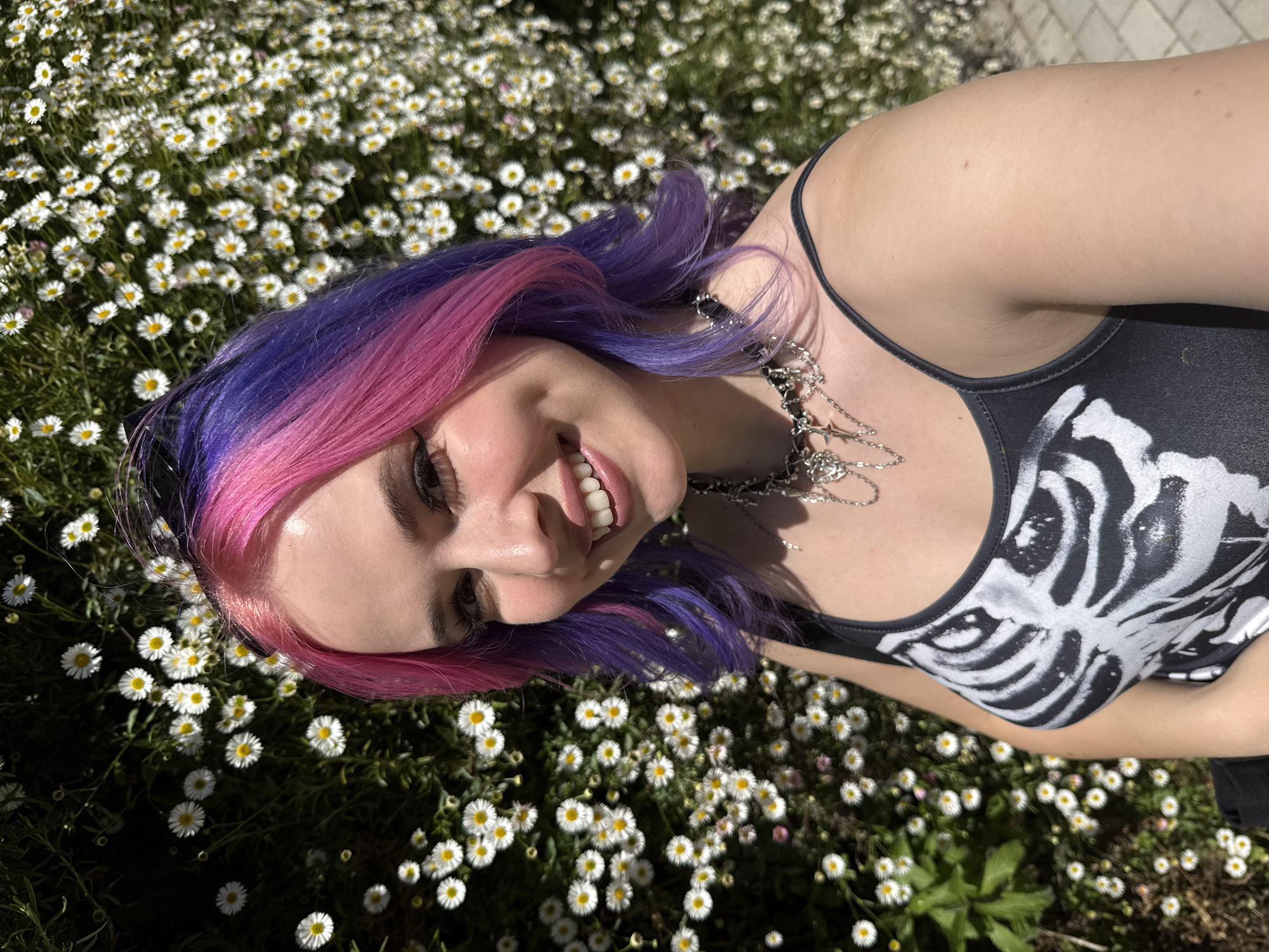 A young woman with pink and purple dyed hair lies on a field of daisies. She is smiling and wearing a black tank top with a white skeleton ribcage design. A silver chain necklace is around her neck. The photo appears to be taken outside on a sunny day. The location is not identifiable as Madrid, Spain, from the visual information in the image.