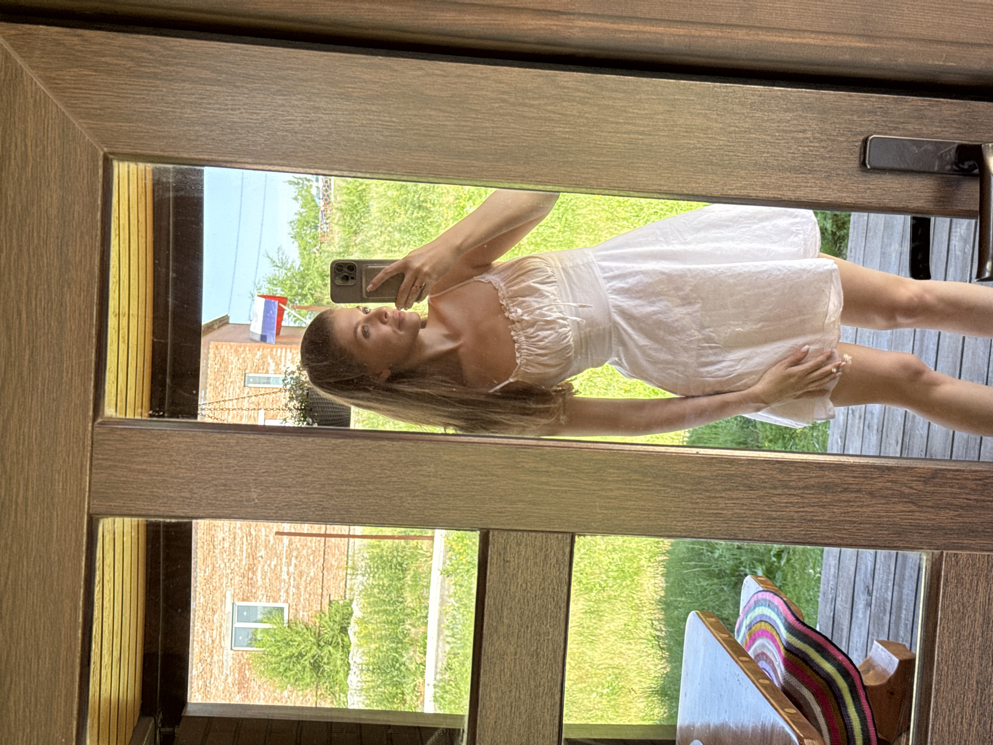 A young woman is taking a selfie in a mirror outdoors. She is wearing a white sundress and has her hair pulled back in a ponytail. She is holding a smartphone in her right hand. The mirror reflects a brick building, a green grassy area, and a blue sky with a flag. There is a wooden table and bench to the left of the frame, and a wooden door with a handle on the right. The scene appears to be in a rural or suburban area.