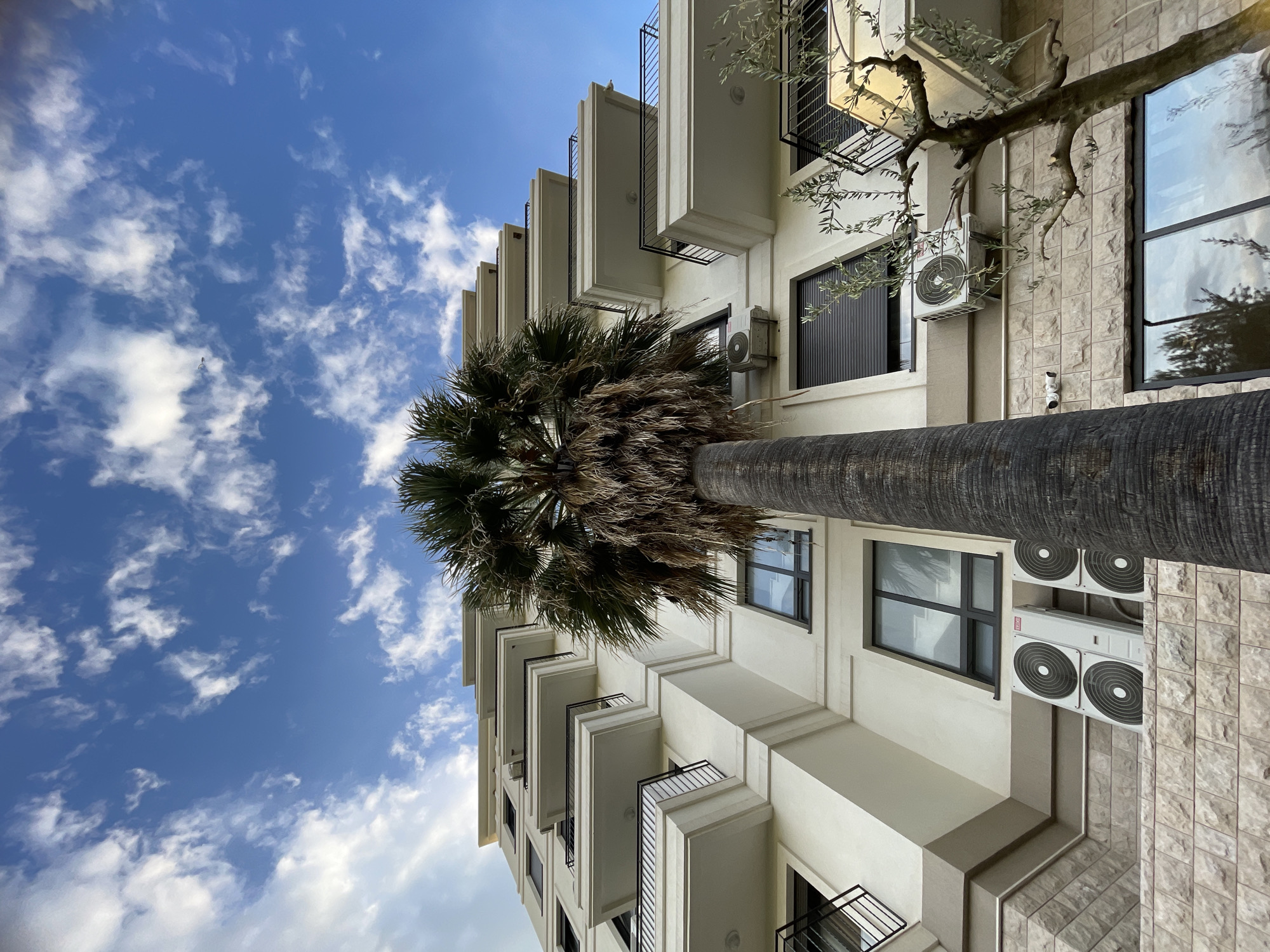 A low-angle shot depicts a multi-story building against a backdrop of a blue sky with scattered white clouds. The building is light-colored with a combination of smooth stucco and textured stone cladding on the exterior. Multiple balconies with dark metal railings jut out from the building's facade. Windows with dark frames are visible, some reflecting the sky. A large palm tree trunk and foliage extend upwards along the building's facade. Air conditioning units are mounted on the building's exterior, adjacent to the windows. Branches from another tree extend from the right side of the frame, partially obscuring the view of the building. The location is inferred to be Bar, Montenegro. The image is taken from below, angled upwards, emphasizing the height of the building and the surrounding environment.
