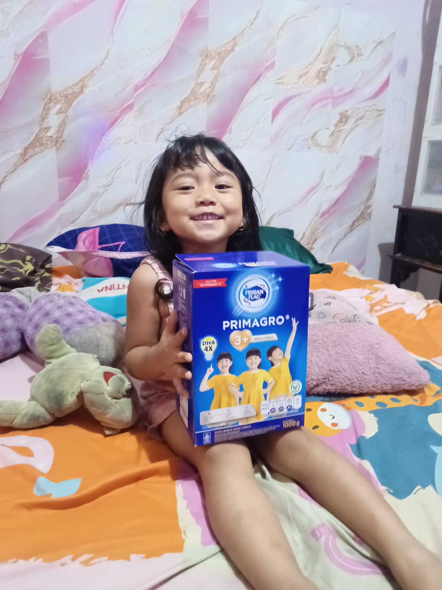 In an indoor setting, a young Indonesian girl with dark hair smiles while holding a blue box of Frisian Flag Primagro milk. She is sitting on a bed with colorful bedding, including orange, green, and teal fabric. To her left, there are several plush toys, including a light green stuffed animal. Behind her, the wall is covered in pink and gold marbled wallpaper. The scene appears to be in a residential area of South Jakarta, Indonesia.