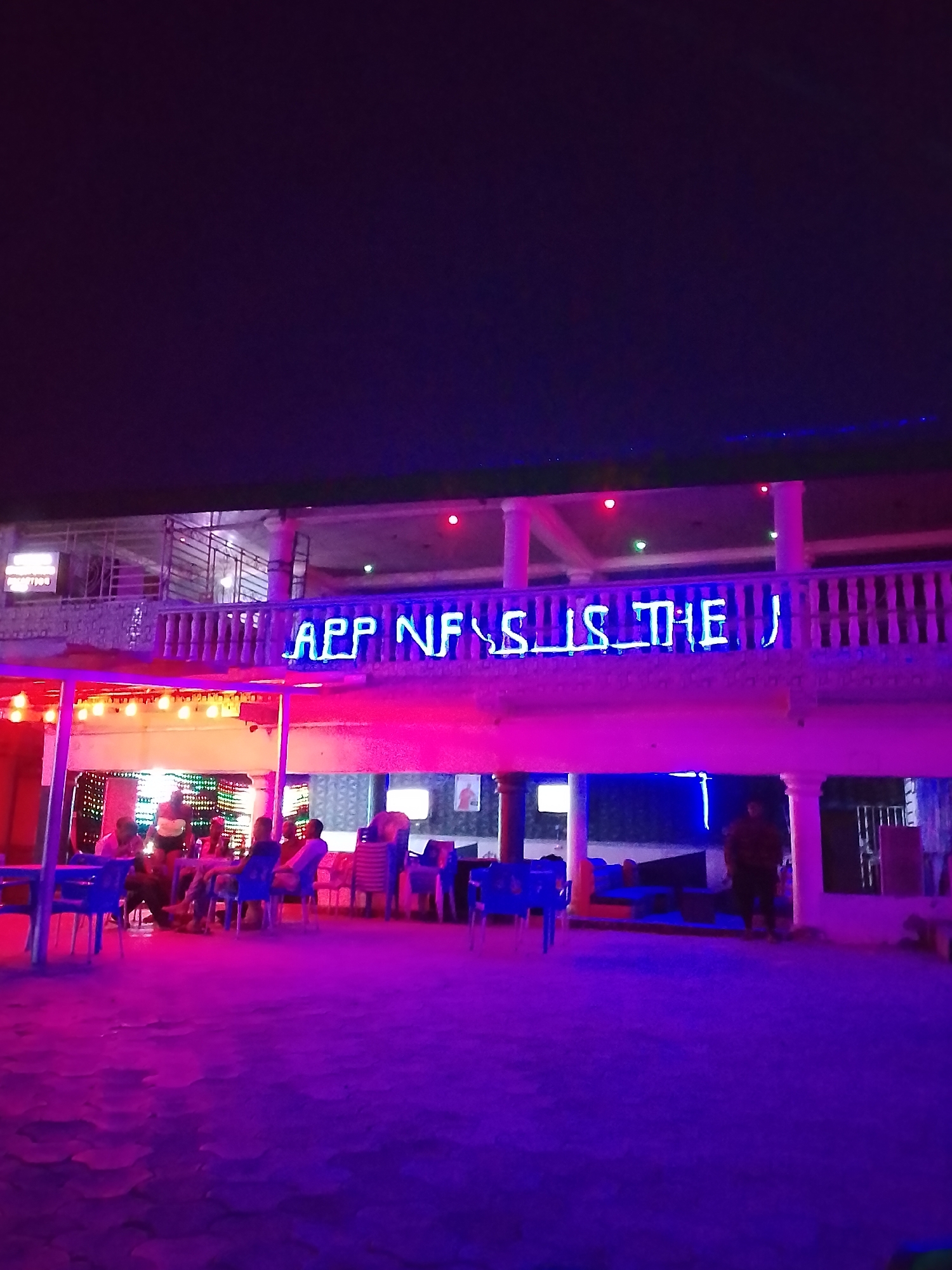 The image depicts an outdoor establishment in Sabon Tasha, Nigeria, at night. Patrons are seated at tables under a brightly lit open-air structure adorned with strands of lights. Above, a two-story building features a balcony with a neon sign that reads "APP NF IS THE U". The ground is a textured surface, possibly pavement or compacted earth, illuminated by pink and purple lighting, giving the entire scene a vibrant, somewhat surreal appearance. A lone individual stands near a building support column. Tables and chairs are present but not all occupied.