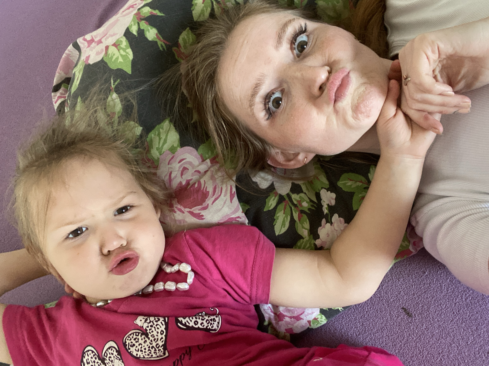 An overhead view captures an adult woman and a young child lying together, looking up at the camera. Both subjects are making playful, "pouty" or "kissing" faces with wide-open eyes, indicating a lighthearted and affectionate moment.
The adult, positioned on the right side of the frame, has light brown hair and fair skin. She wears a light-colored, possibly off-white or pale grey, top. Her left hand is visible near her chin, revealing a ring on one finger.
To her left, the young child, likely a toddler, has light brown hair and fair skin. She is dressed in a vibrant pink t-shirt adorned with leopard-print heart designs and some indistinct cursive text that appears to say "happy day" and "hello". She wears a necklace made of irregular white beads. Her right arm is bent, resting near her head.
They are lying on a dark-colored pillow or cushion featuring a pattern of pink and green flowers and leaves. Beneath them, and extending across the bottom and left side of the image, is a textured purple surface, possibly a carpet or blanket. The scene conveys a warm and playful interaction, set indoors. The location context provided is Chaikovsky, Russia.