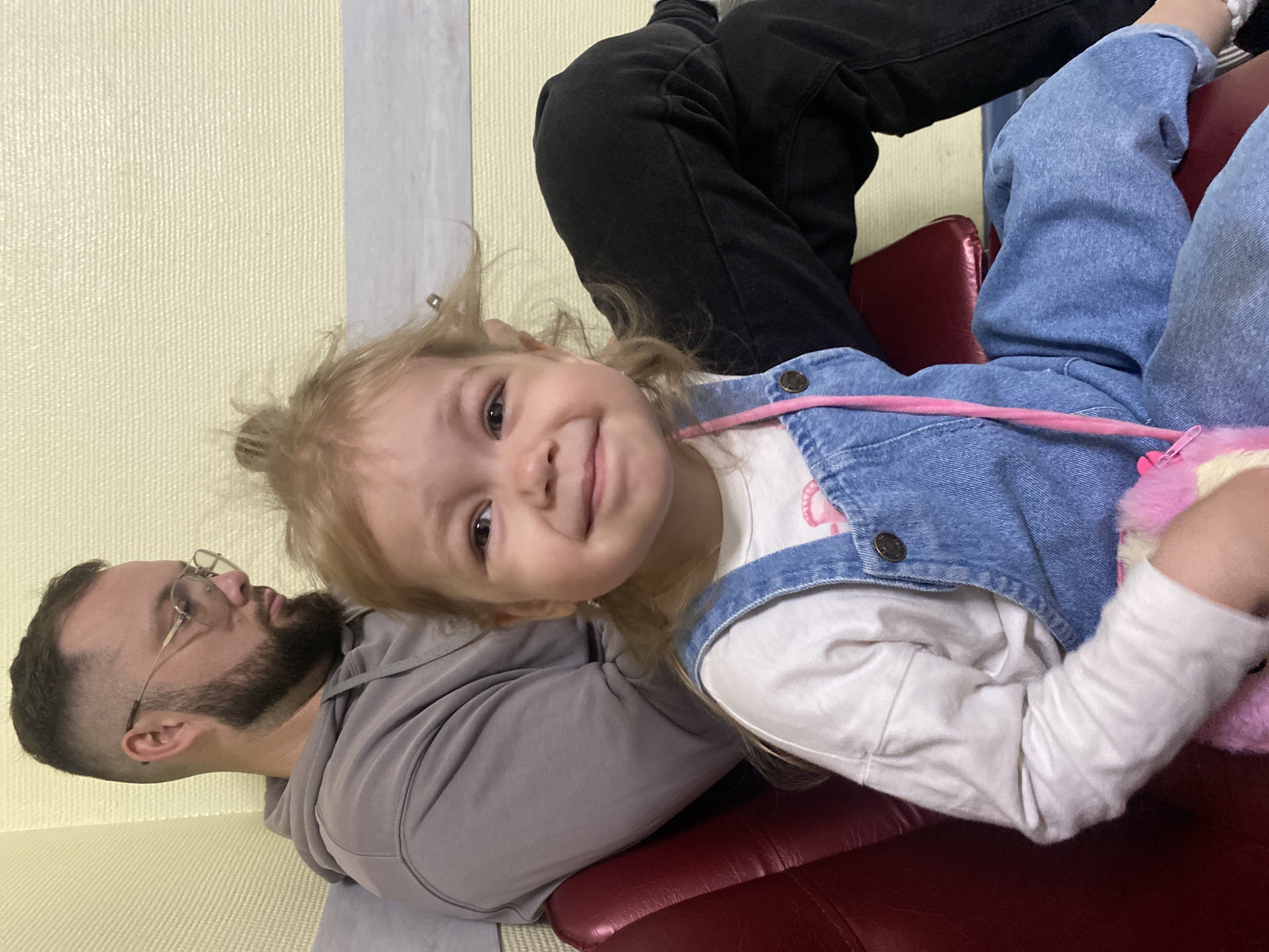 In a room with pale yellow walls, a man and a young girl are seated side-by-side on a red chair. The man, positioned on the left, wears a grey hoodie and glasses, with a beard and a fade haircut. He looks downwards, towards his right. The girl, in the foreground and to the right, is wearing denim overalls over a white long-sleeved shirt and a pink accessory hanging from her overalls. Her hair is pulled up messily. She is looking directly at the camera with a slight smile. The environment appears to be indoors, potentially a waiting area or a room in a residence. There is no clear indication of the city of Chaikovsky, Russia, in the image.