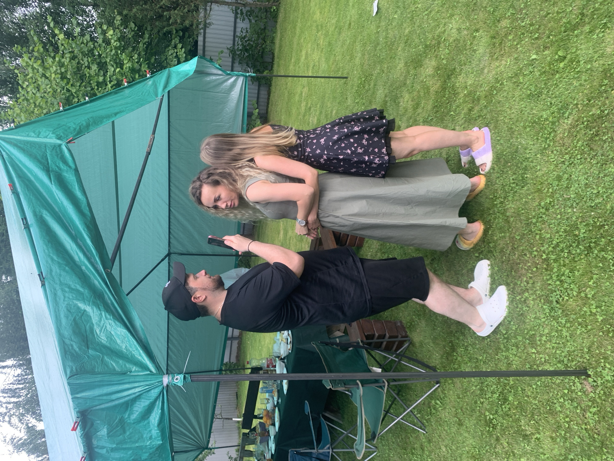 In Sergeevo, Russia, a man in a black t-shirt and shorts holds a smartphone, seemingly recording or taking a picture of two women. The woman in the foreground, wearing a long olive-green dress, stands with her arm around the woman next to her, who is in a black floral dress. They are standing on a grassy lawn under a green tarp canopy. A table laden with food and drinks is visible to the left, along with several folding chairs. A wooden fence and trees form the background. The ground is covered in green grass with some clover.