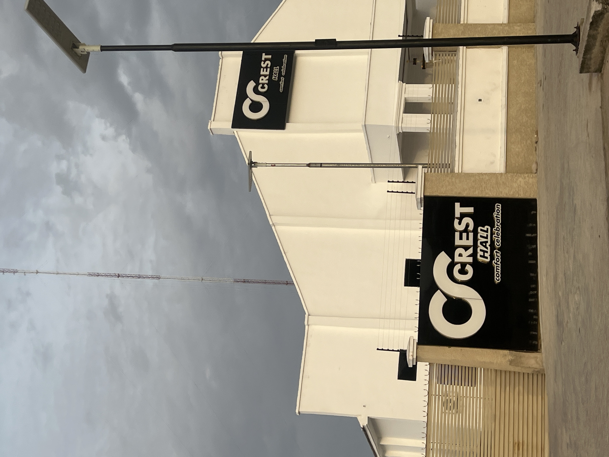 The image shows a building with the sign "Crest Hall comfort celebration," along with a streetlight. The building appears to be located in Howayi, Nigeria, based on the sign. The sky is overcast. A tall antenna or communications tower is visible in the background. The building is white with architectural details like railings and ledges. The photo is taken at an angle, and there is a concrete sidewalk or roadway in the foreground.