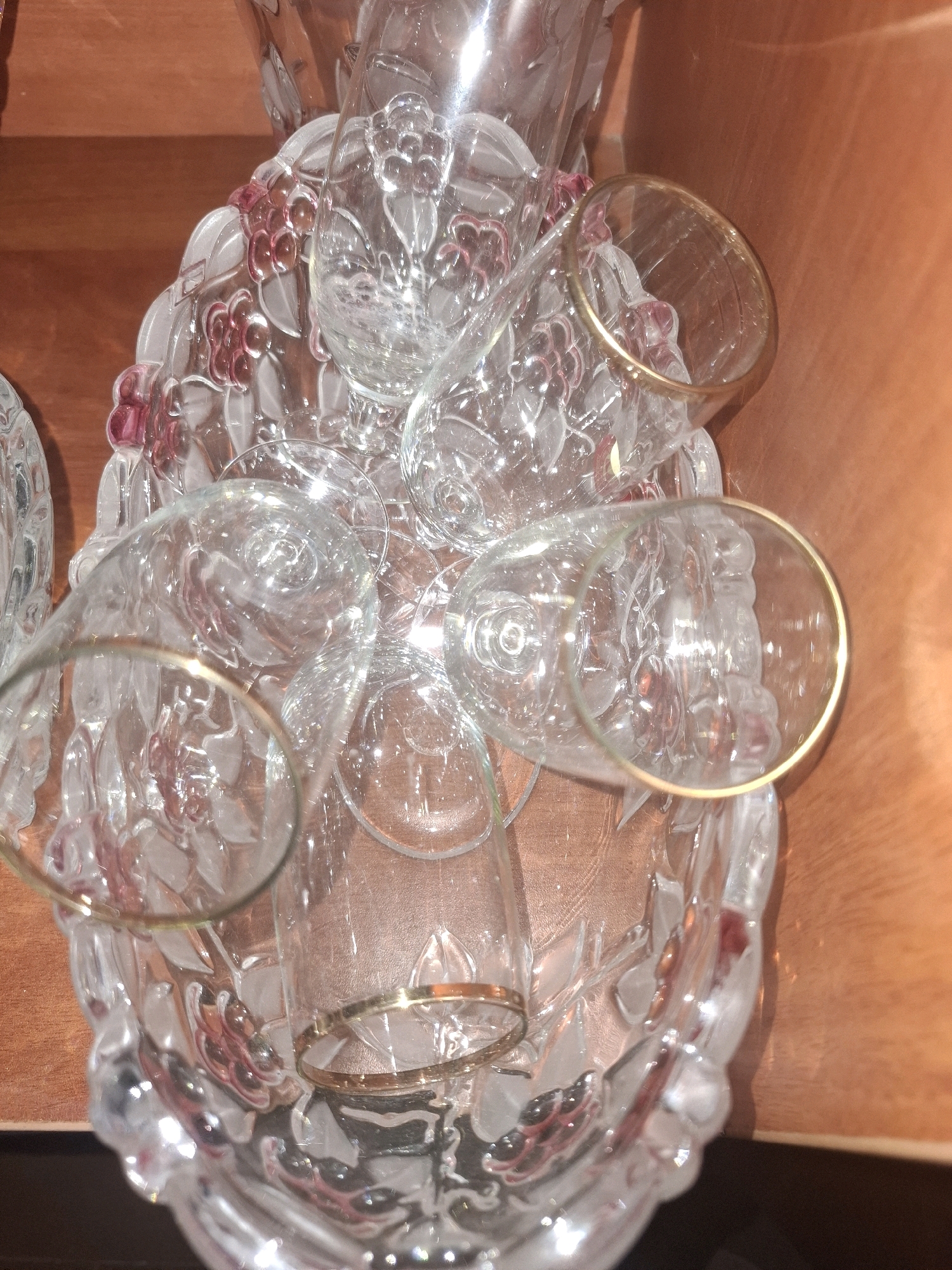 A close-up, high-angle shot captures a collection of clear glass stemware, including wine glasses and possibly a decanter, arranged within a decorative glass bowl. The bowl itself is intricately patterned with what appear to be grapes and leaves, some of which are tinted a deep red or pink. The rims of the wine glasses are adorned with a thin gold band. The stemware is clustered together, suggesting they are being stored or presented. The background consists of a textured, light brown surface, possibly a wooden table or shelf. The lighting is warm and creates reflections on the glass surfaces. There are no people visible in the image, and the scene is static. The location is unknown and there is no information to suggest it is in Almaty, Kazakhstan.